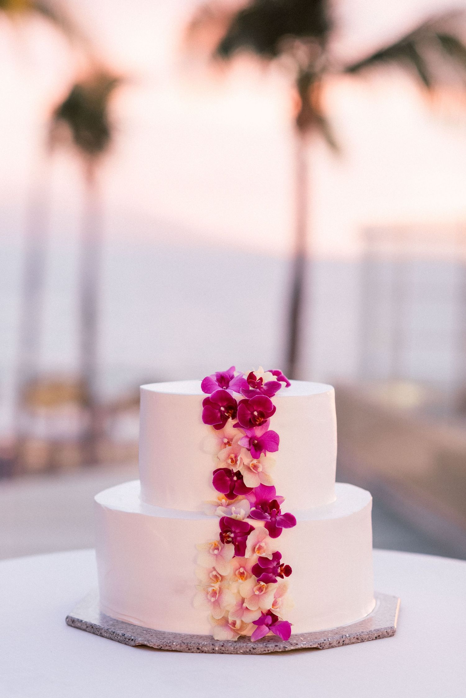 Two-tier white wedding cake decorated with cascading purple orchids against palm tree backdrop.