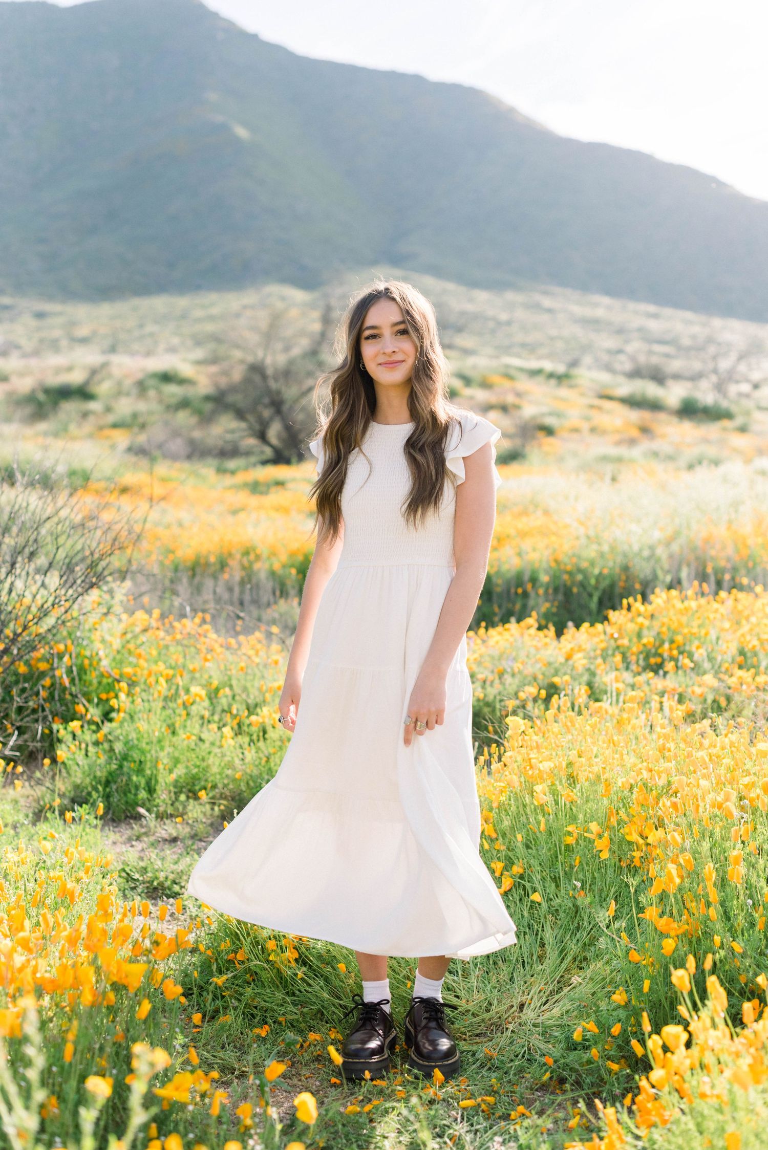 White flowing dress contrasts with yellow wildflowers and mountain landscape.