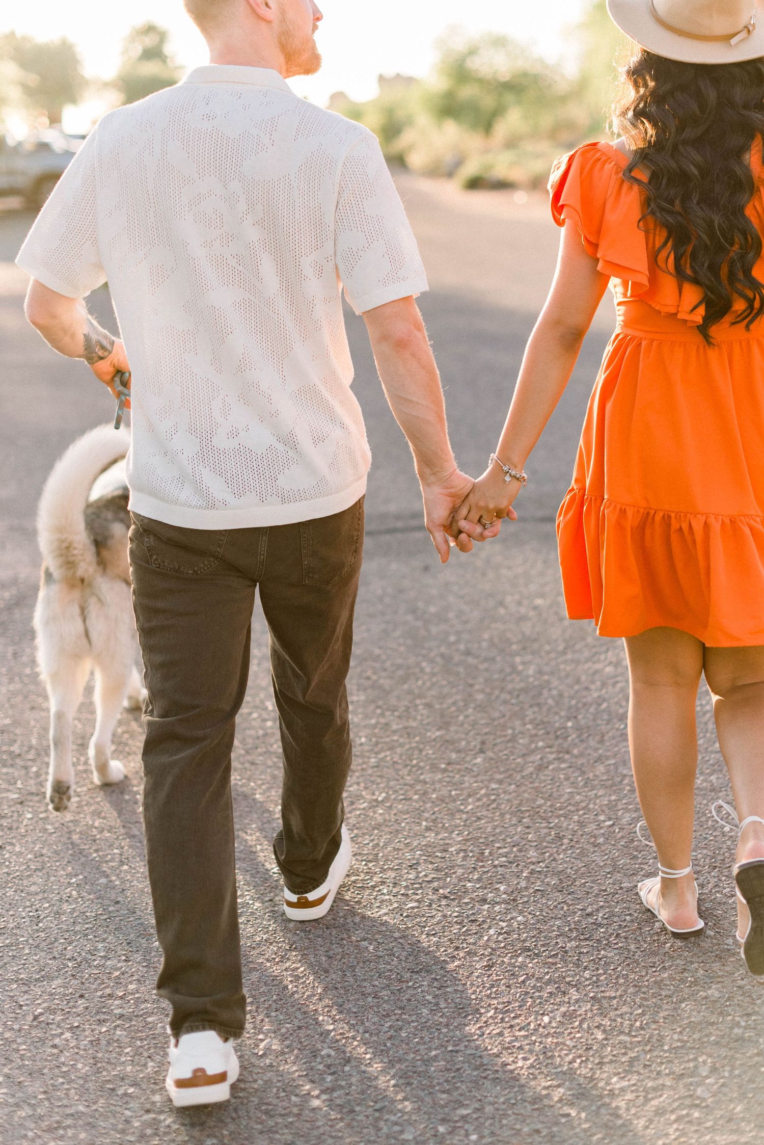 Back view of couple walking hand in hand with their dog on desert path at sunset.