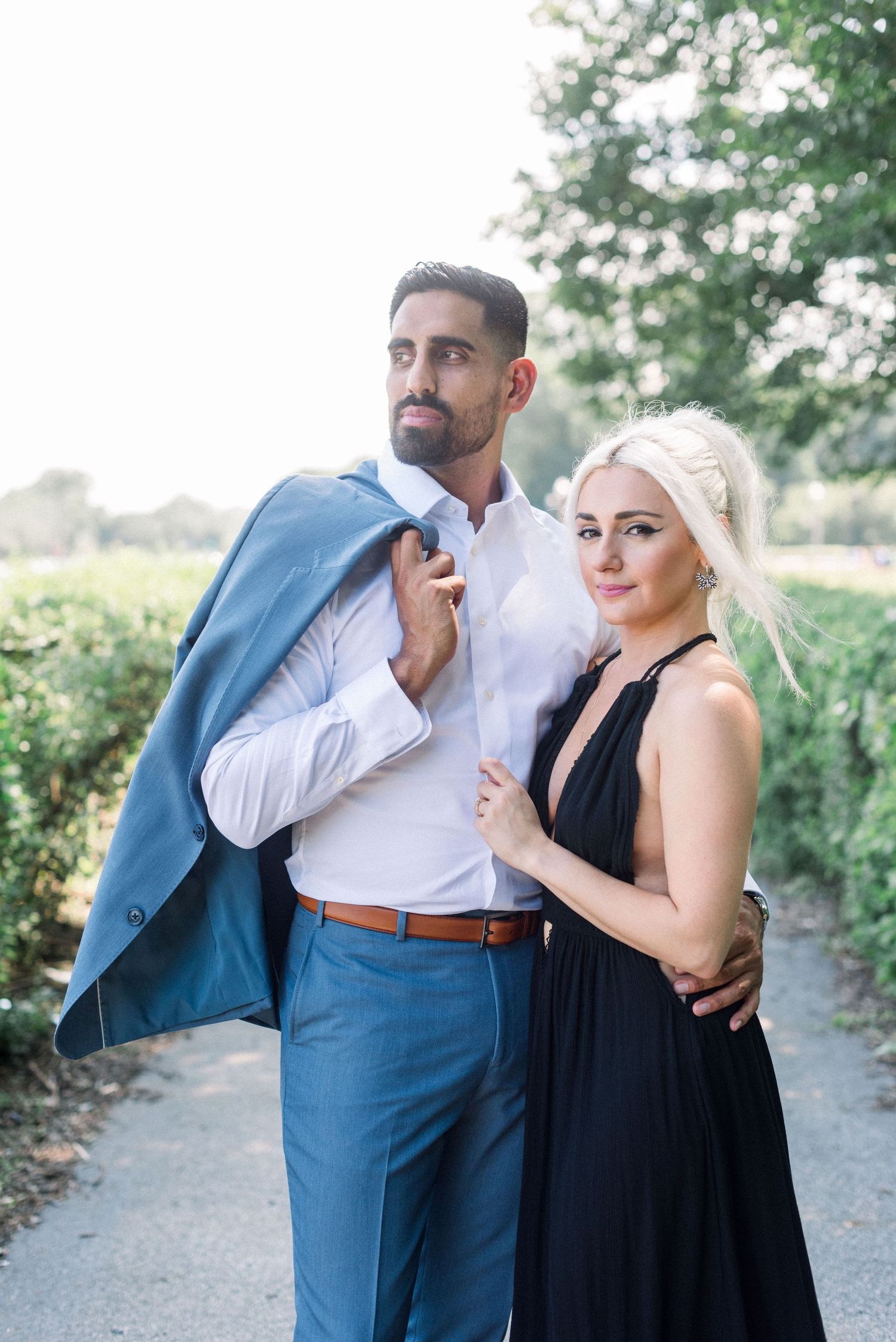 Elegant couple in formal wear posing together on garden pathway with man holding blue jacket.