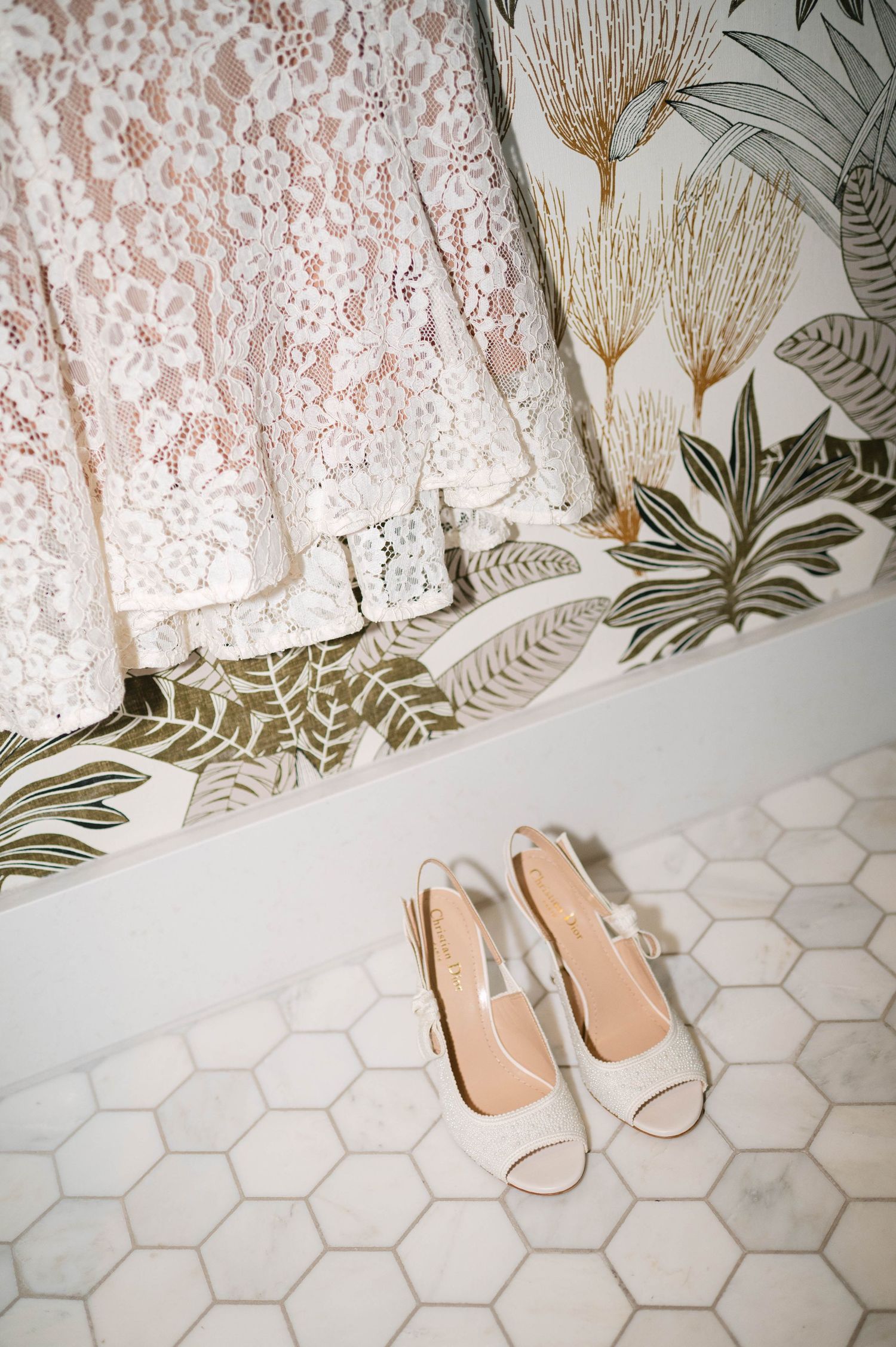 White lace dress hangs against floral wallpaper with blush pink sandals on hexagonal tile floor.