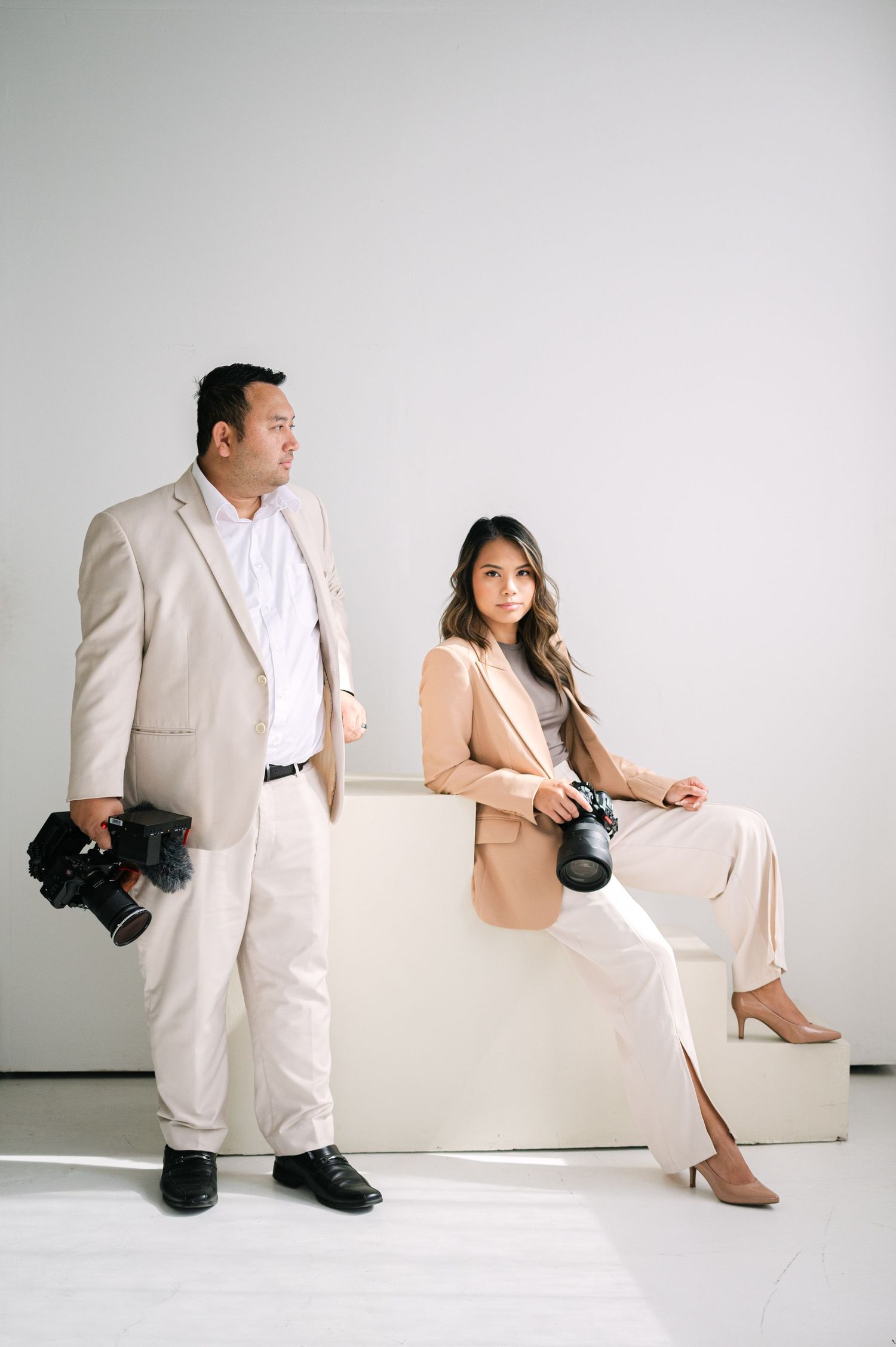 Two photographers in matching beige and white outfits pose with professional cameras against a white studio backdrop.