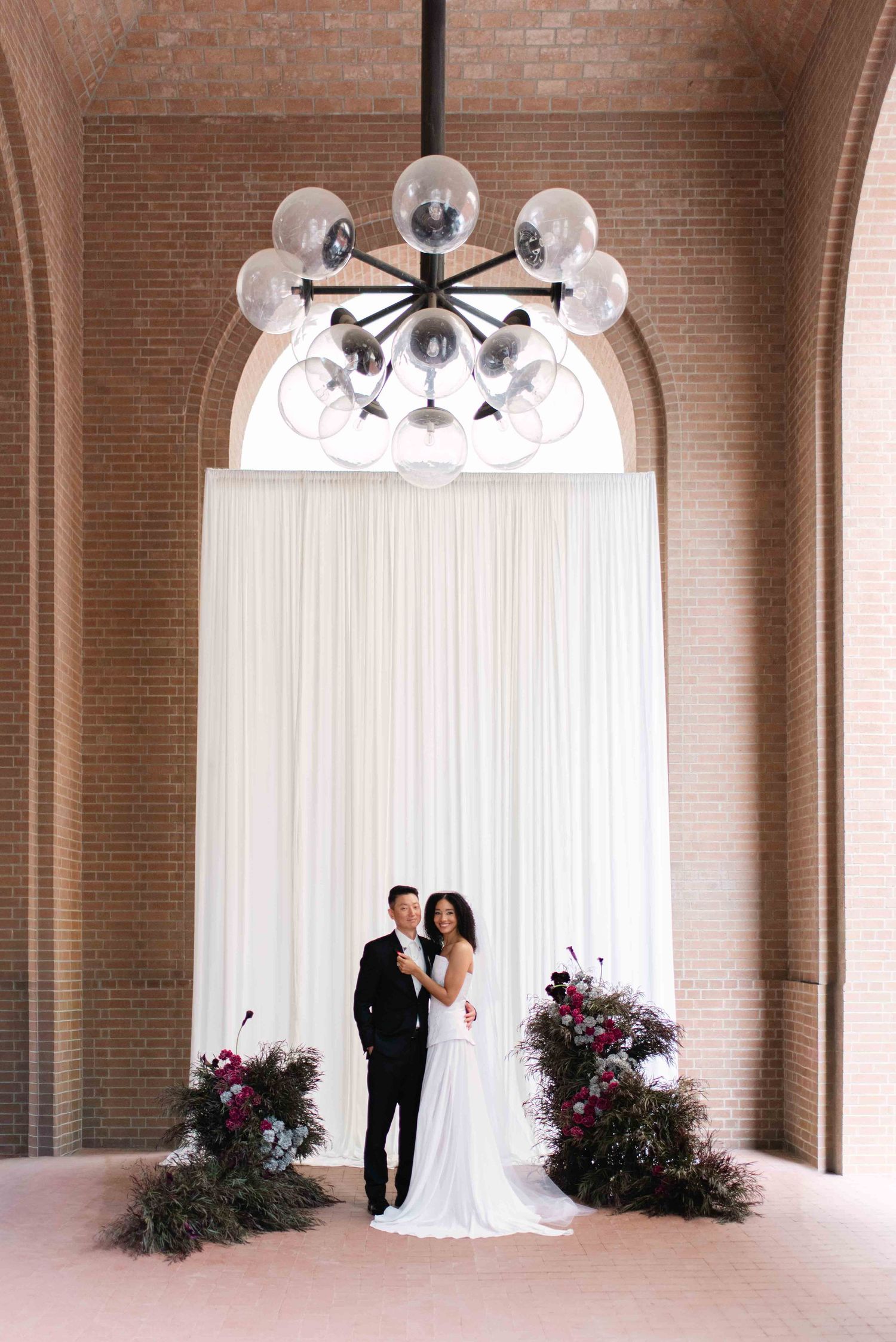 A romantic wedding couple stands in an elegant brick archway with white chandeliers and pink floral arrangements.