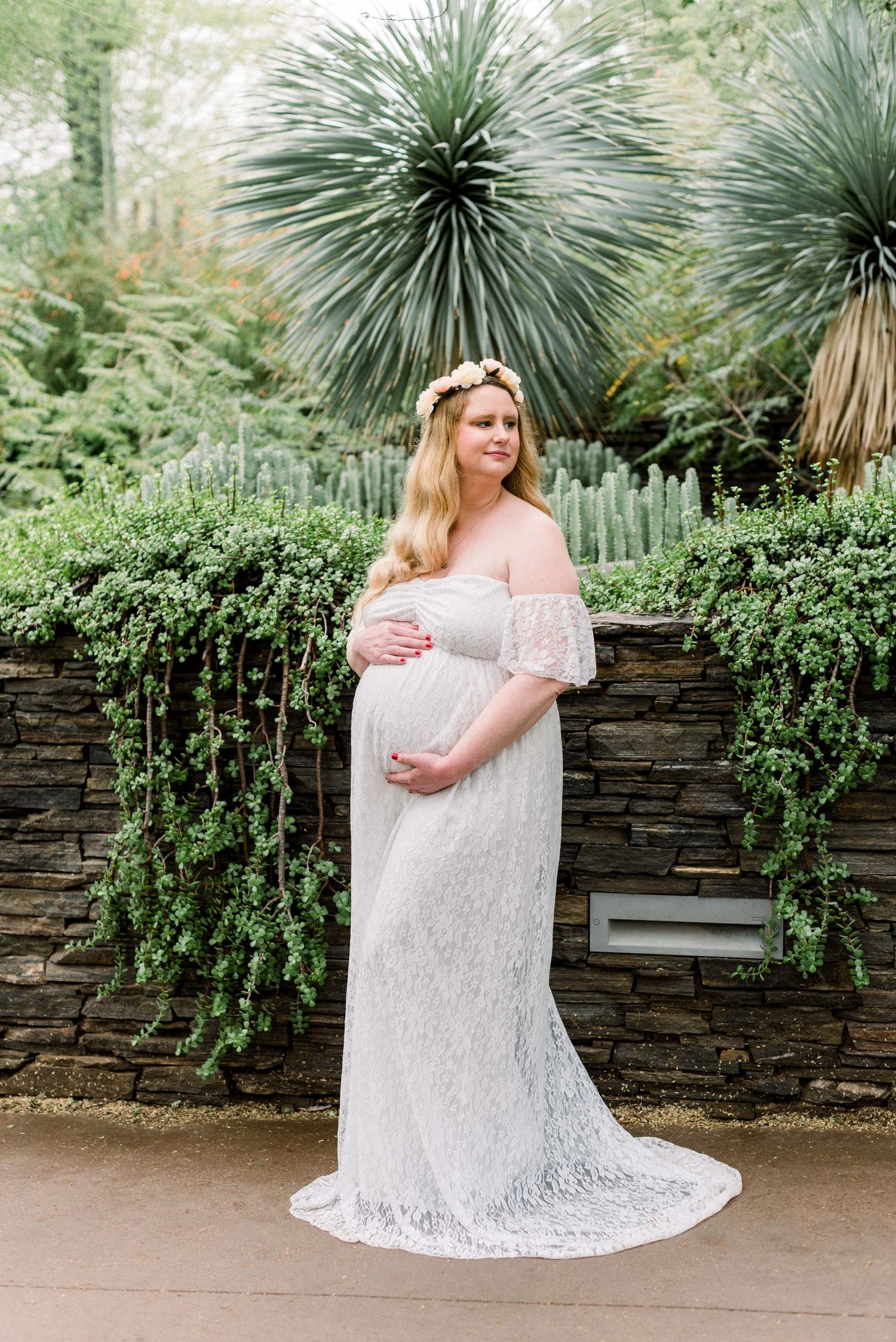 Maternity portrait in a white lace gown standing in front of a stone wall with tropical plants and yucca trees.