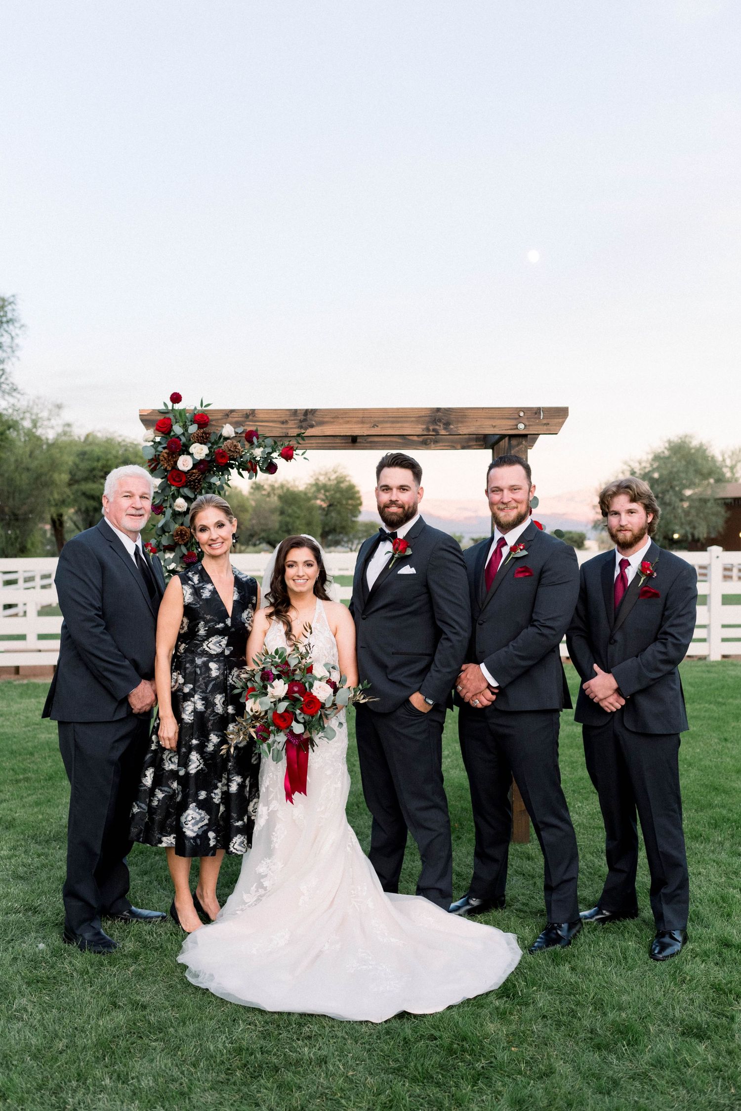 Wedding party poses under wooden arch decorated with red roses outdoors.