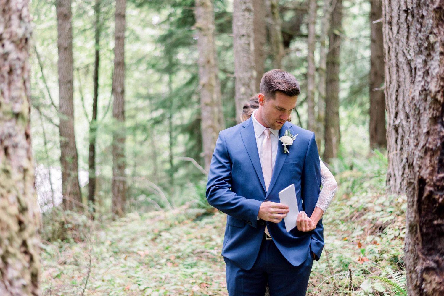 Groom reading a letter while standing in a serene forest setting.