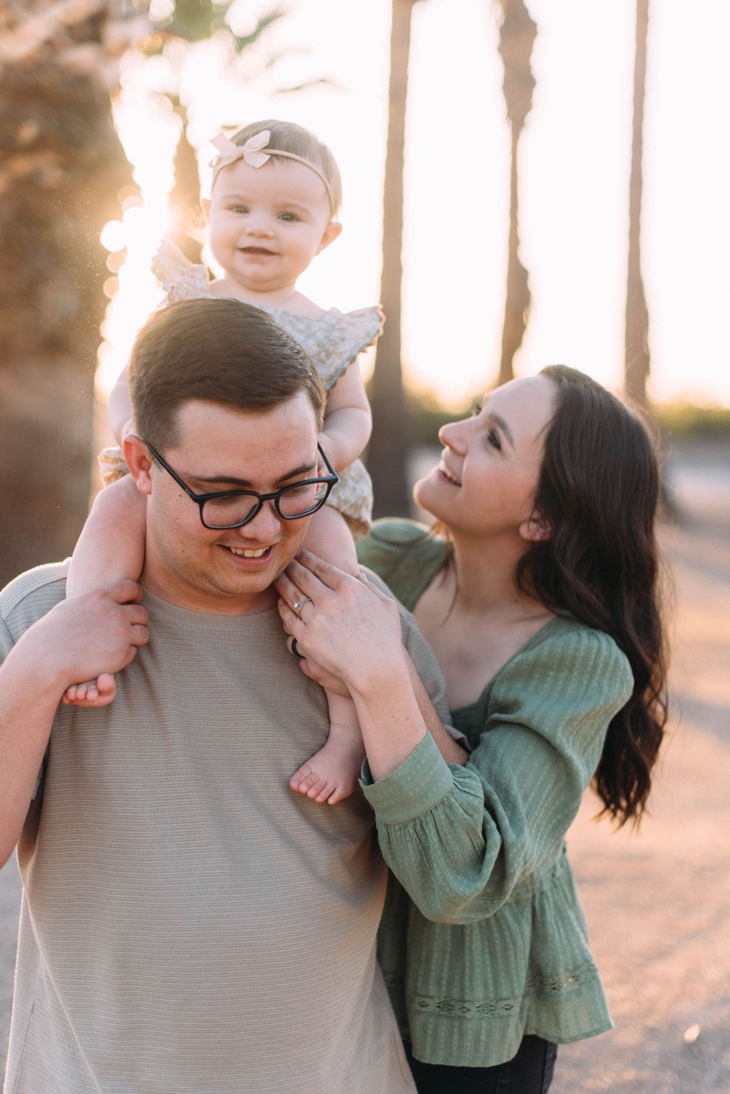 Family enjoying sunset moment with baby on shoulders near palm trees.