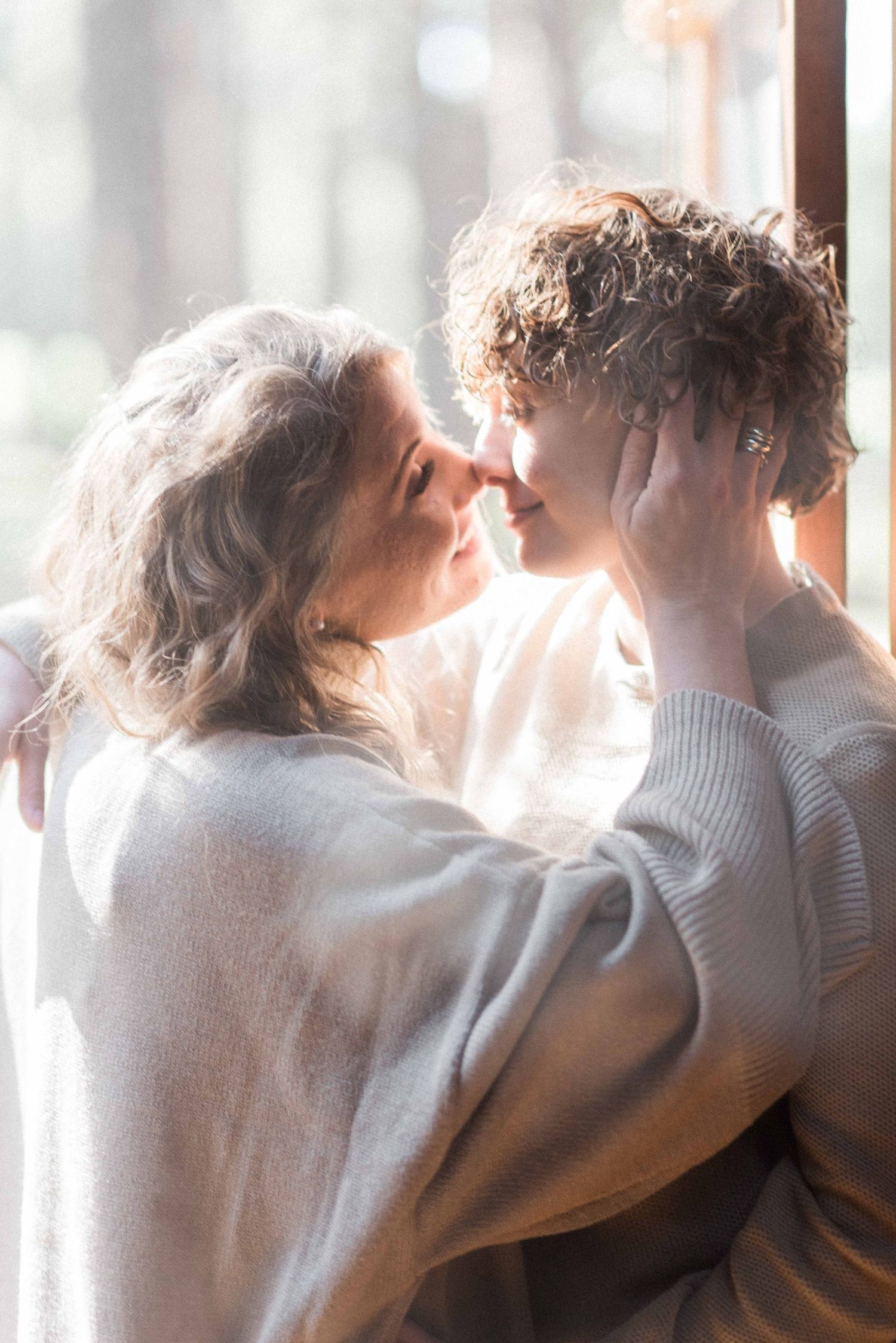 Intimate couple in cozy sweaters sharing tender moment by window in soft lighting.