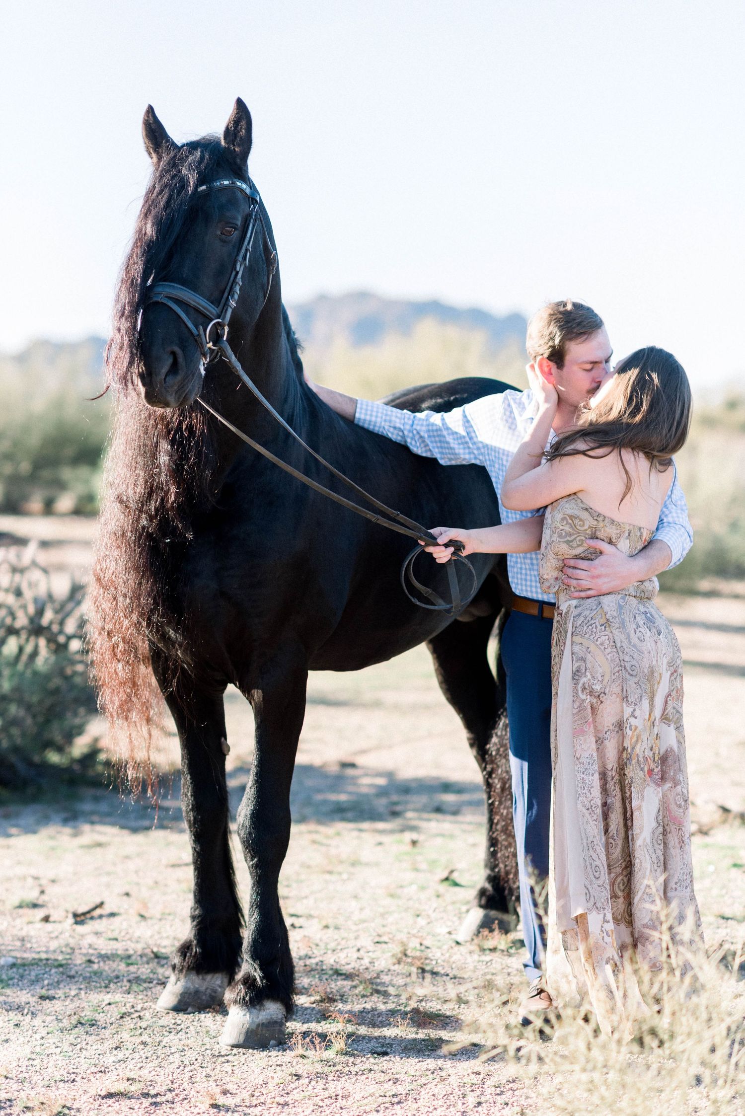 Couple shares romantic kiss next to black horse in desert landscape.