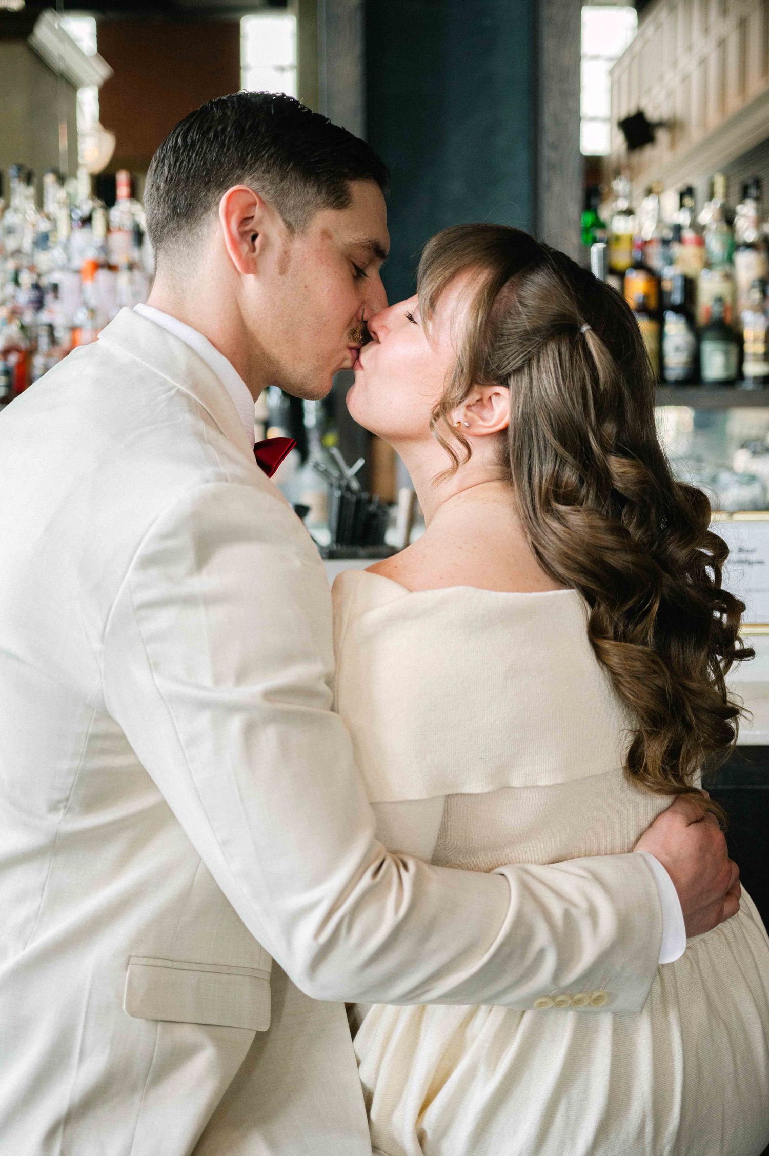 A tender embrace between a couple in cream-colored formal wear against a bar backdrop.