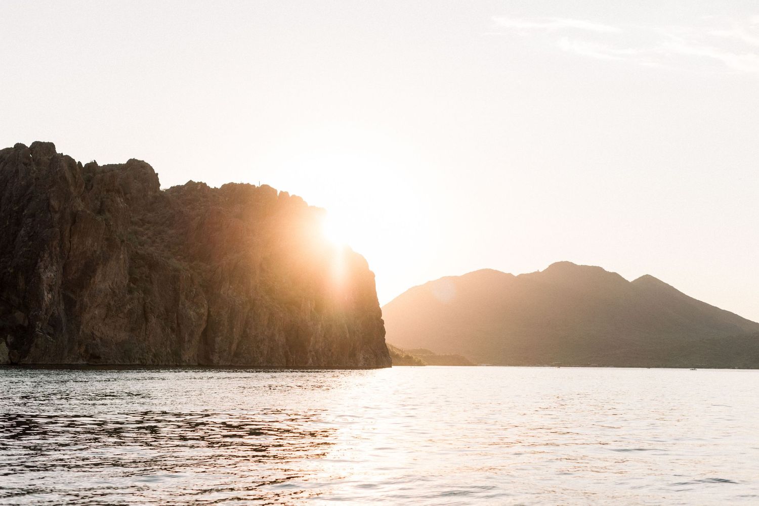 Dramatic sunset over mountain cliffs and lake with orange sunlight peeking through.