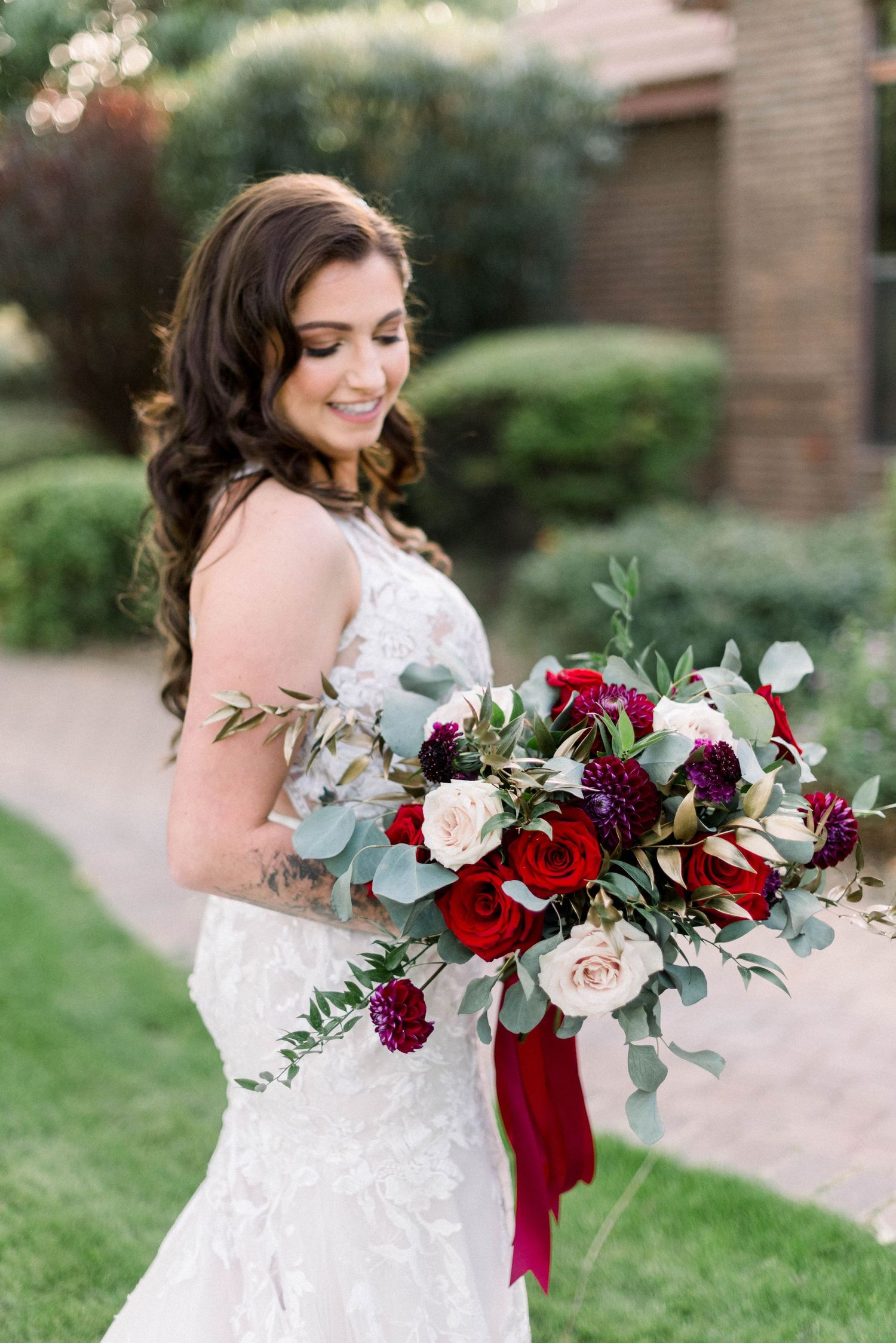 Side profile of bride holding romantic bouquet with red roses and trailing burgundy ribbon.
