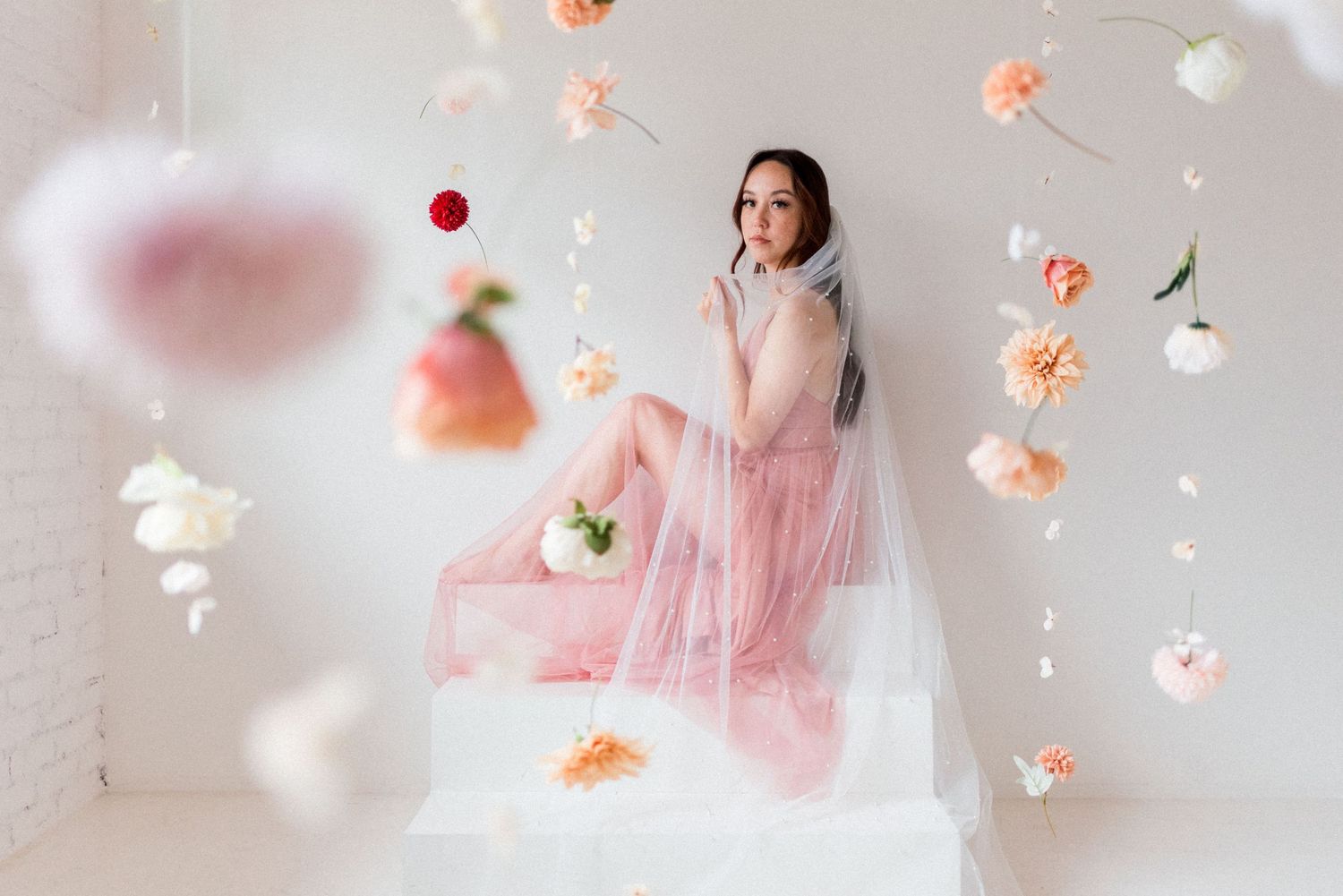 Whimsical portrait with floating peach flowers and pink tulle dress in white room.