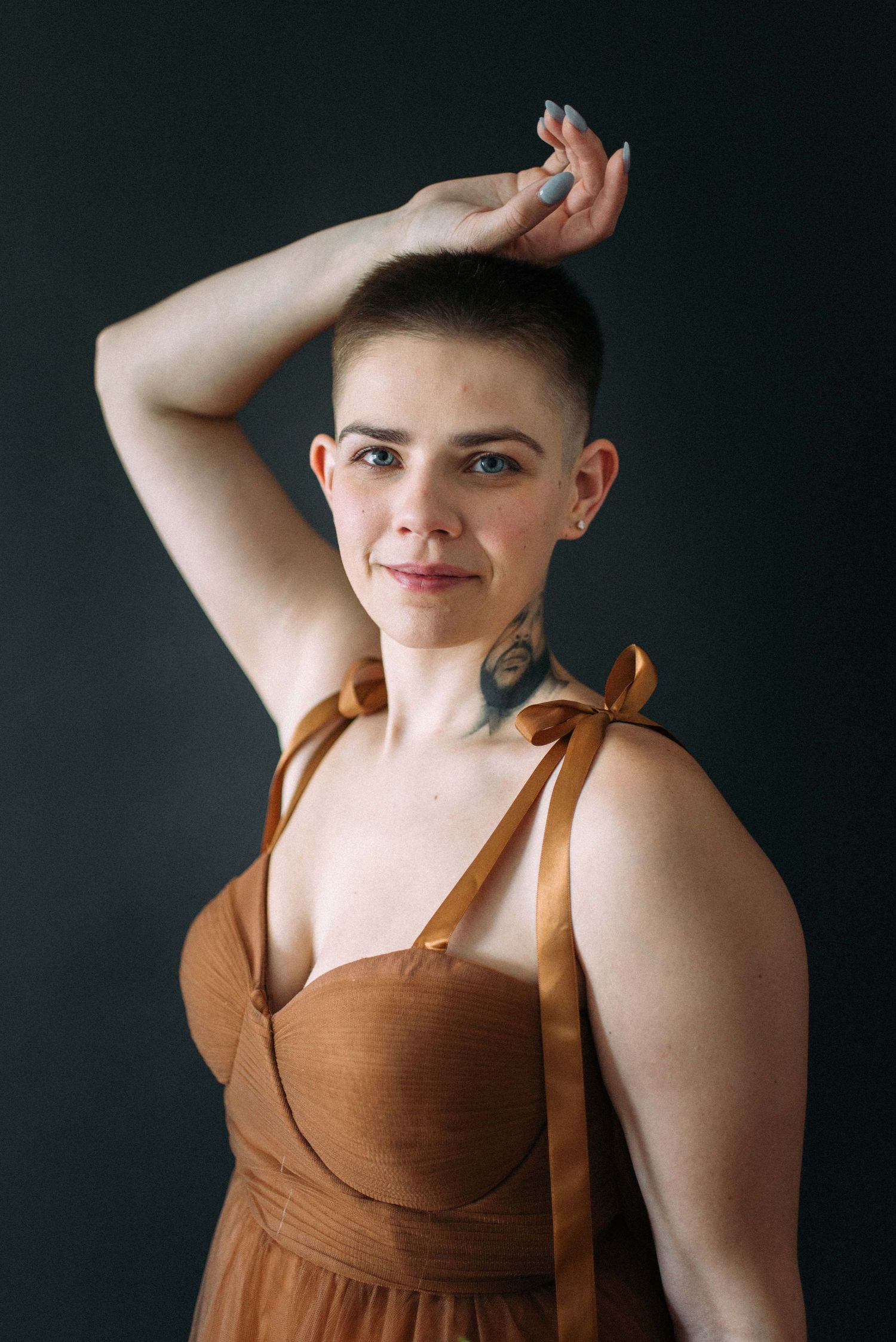 A graceful studio pose against a dark background wearing a brown sleeveless dress.
