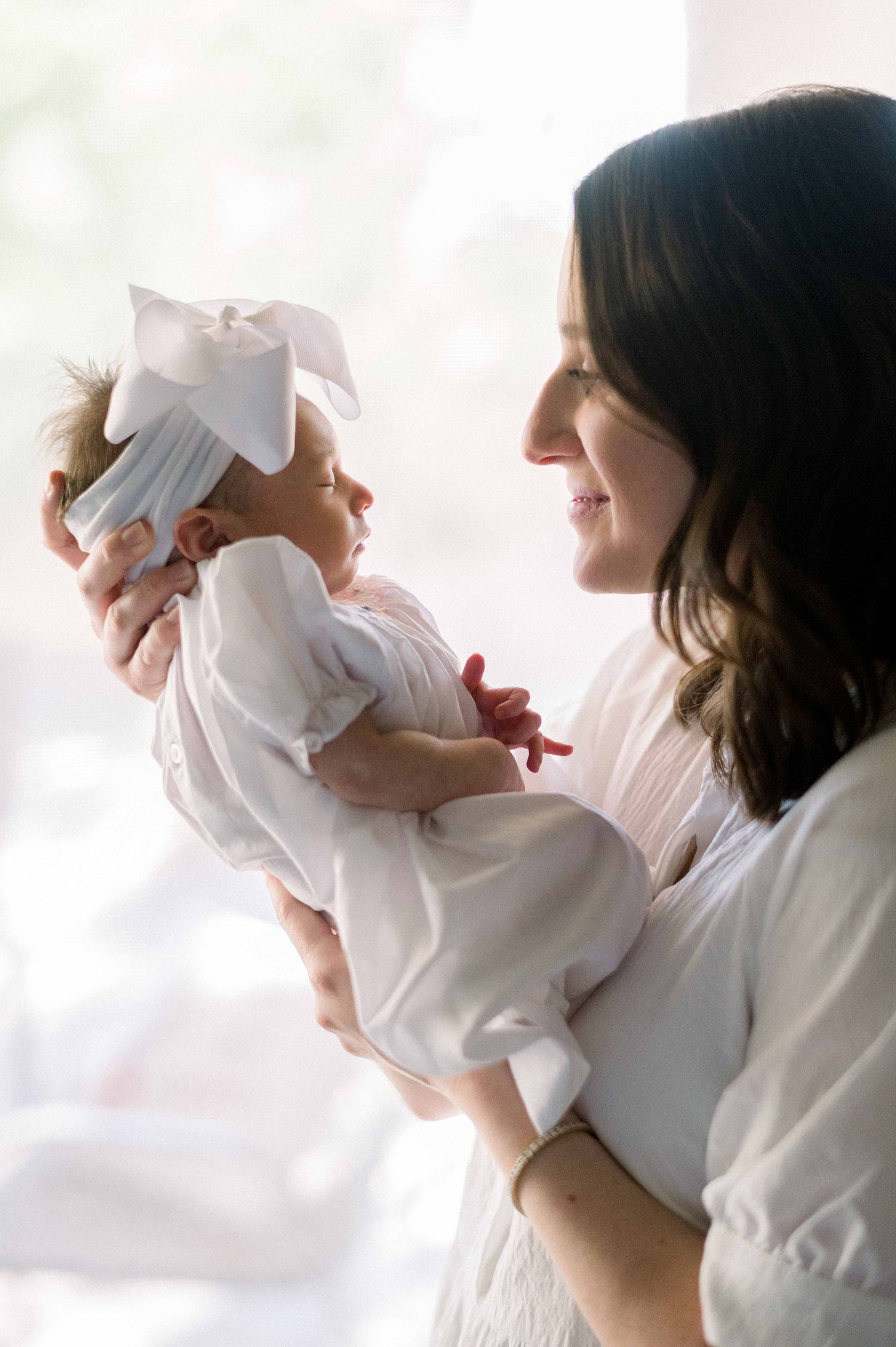 A woman in white cradles a baby wearing a bow while standing in soft natural light.