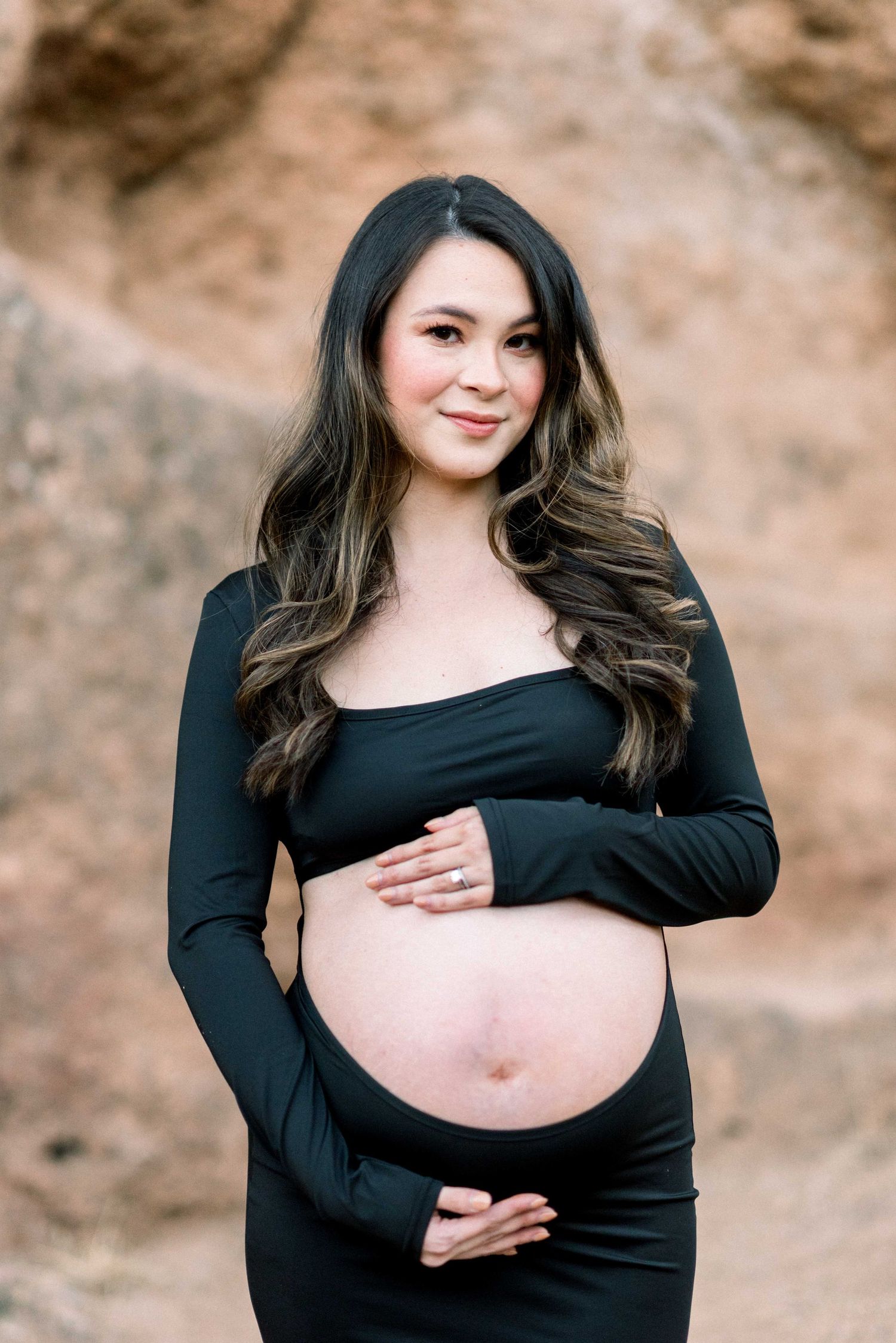 Maternity portrait of a woman in black dress showing her pregnant belly against a rocky outdoor backdrop.