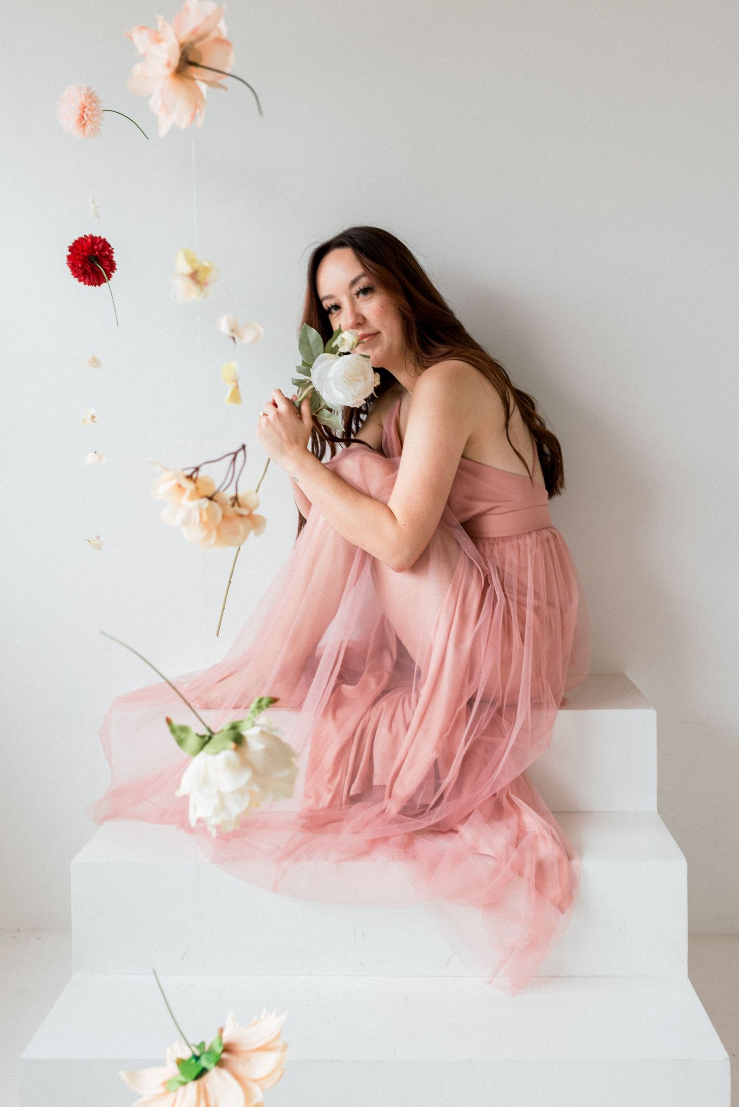 Romantic portrait with pink tulle dress on white steps surrounded by suspended flowers.