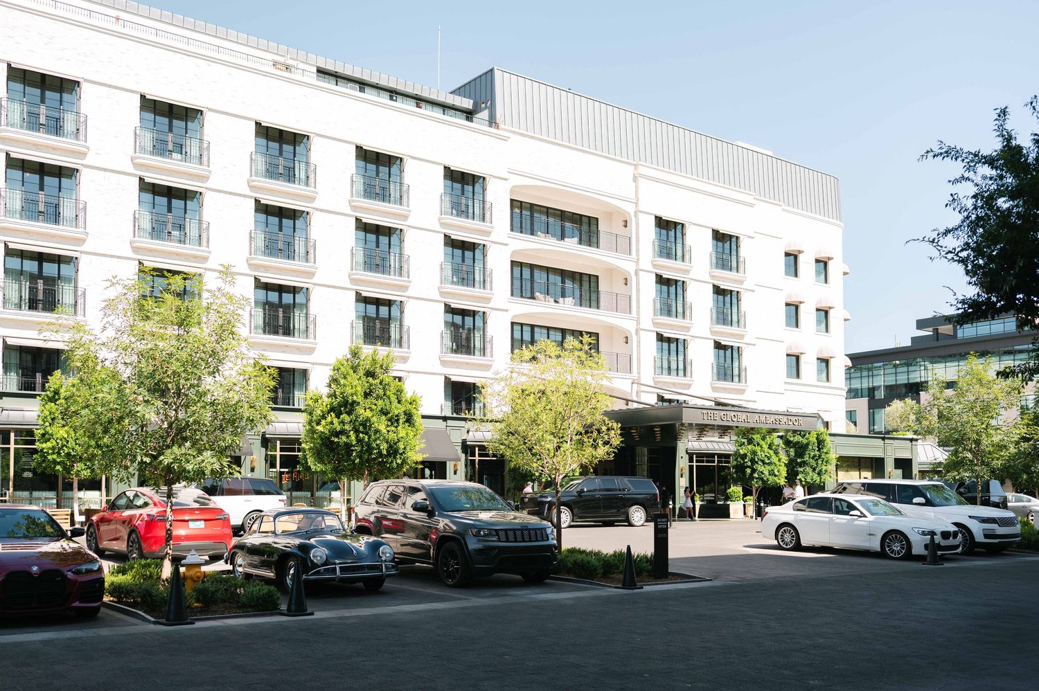 Modern hotel building exterior with white facade, balconies, and parked cars in front parking lot on sunny day.