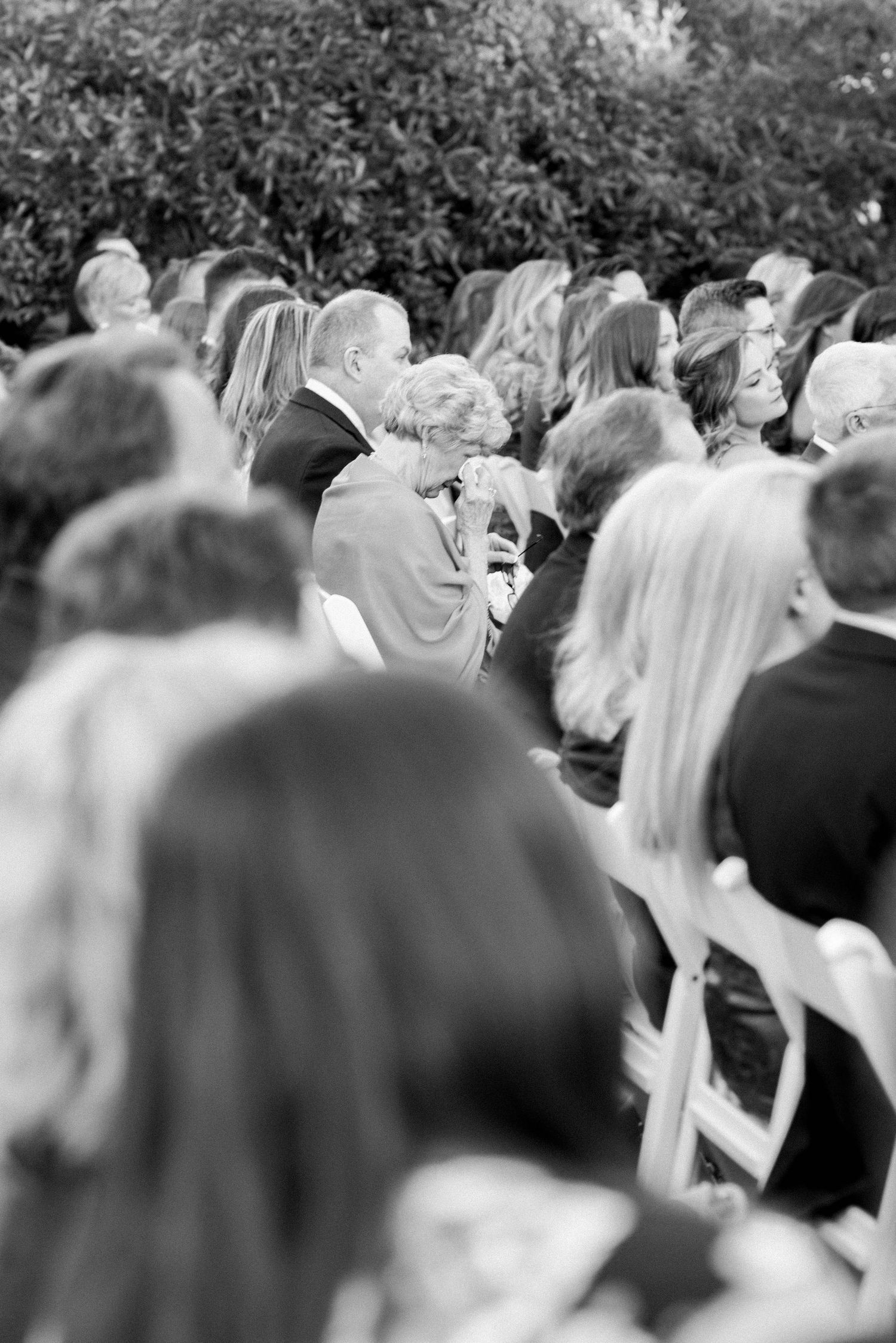 A black and white photograph shows a crowd gathered at an outdoor event with people seated in rows of chairs.
