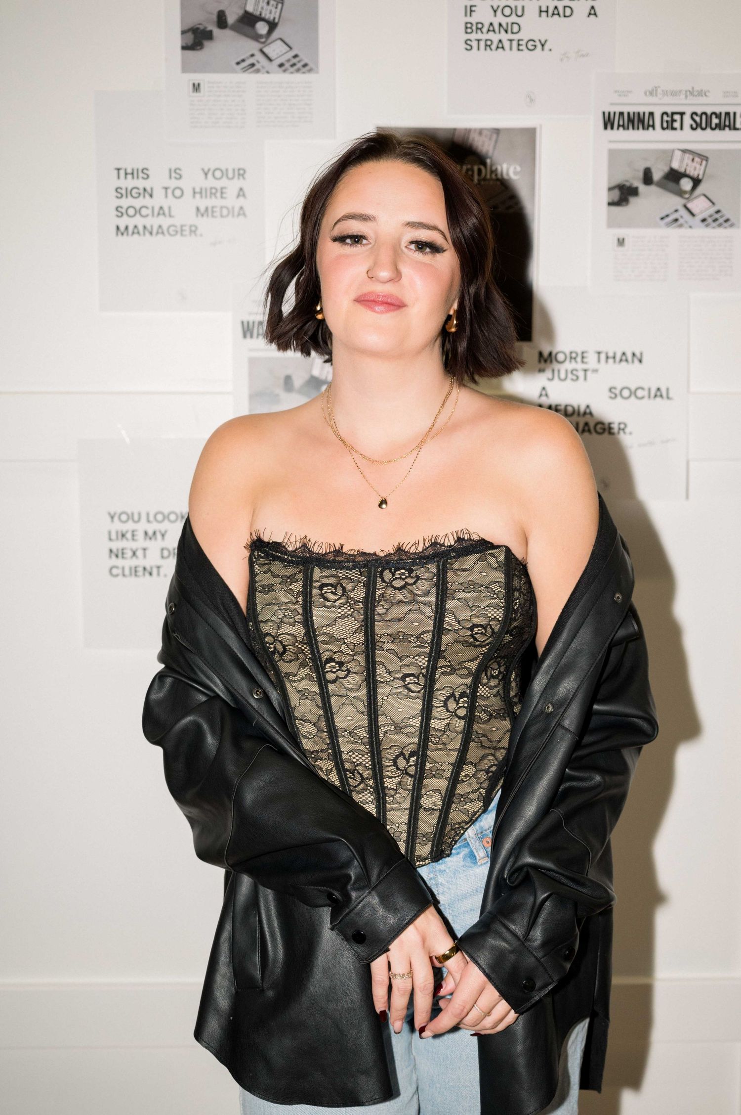 A portrait against a white wall wearing a black leather jacket and corset top.