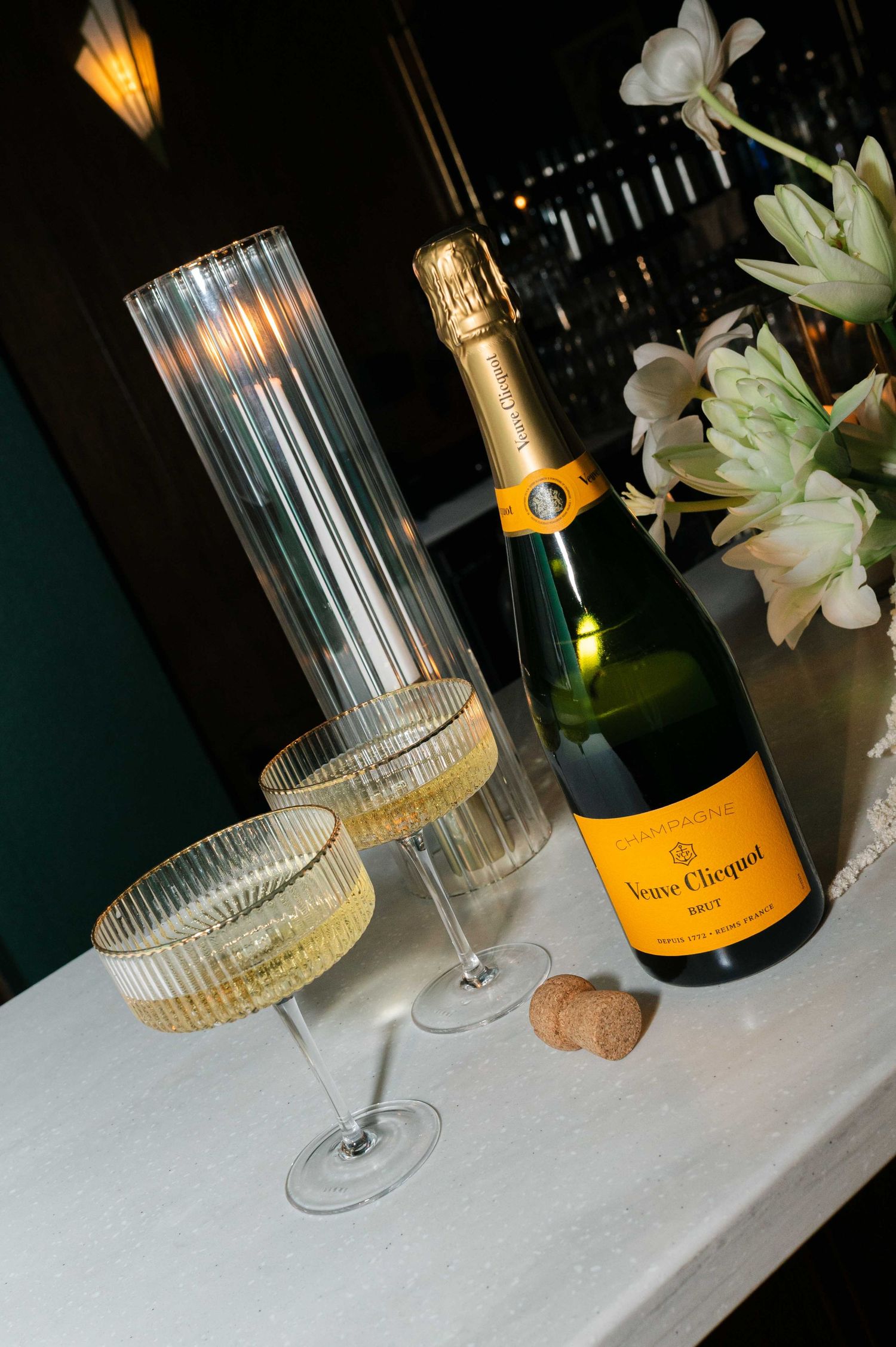 Elegant table setting with Veuve Clicquot champagne, crystal glasses, and white flowers in dark ambient lighting.