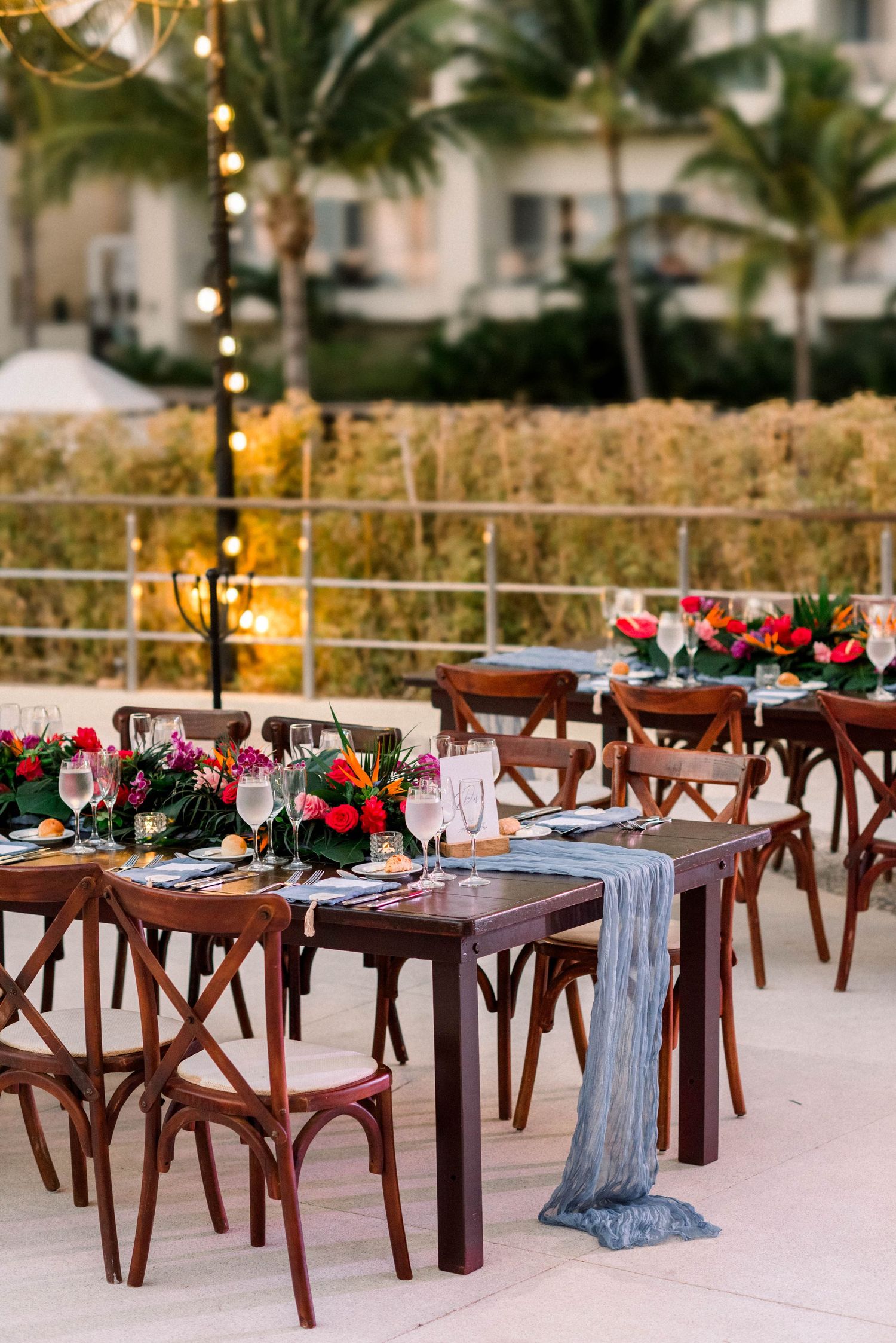 Romantic outdoor dining setup with wooden chairs and floral centerpieces under palm trees and string lights.