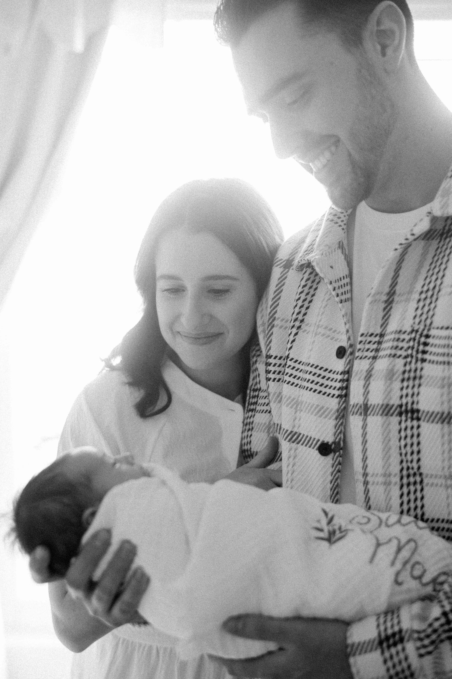 Intimate black and white portrait of parents lovingly holding their newborn baby in natural light.