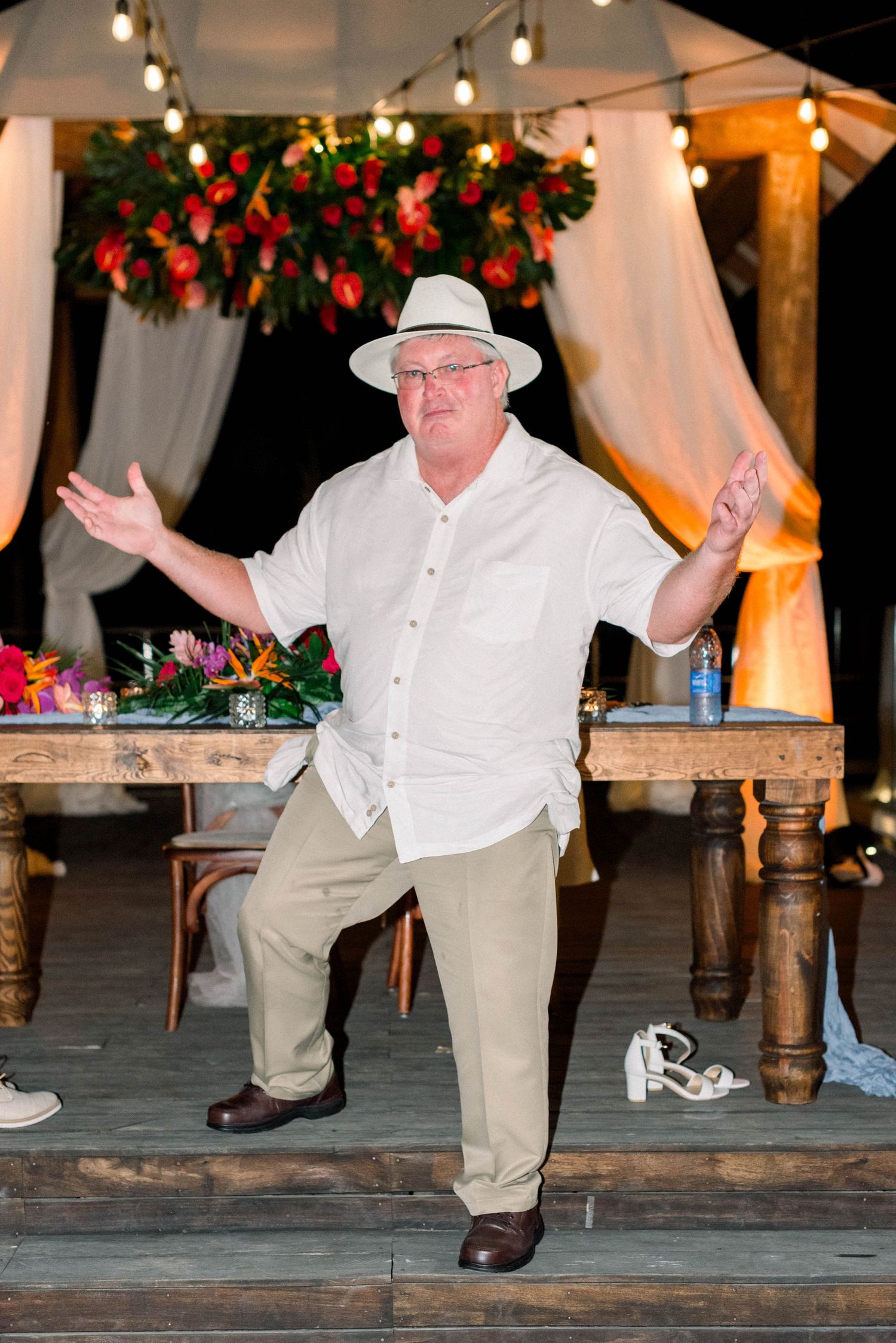 Wedding guest in white hat dances enthusiastically on decorated outdoor stage.