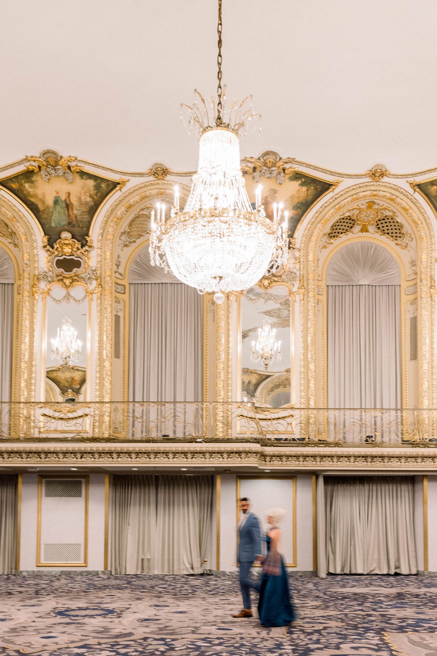 Blurred figures in formal attire stand beneath crystal chandelier in ornate ballroom.