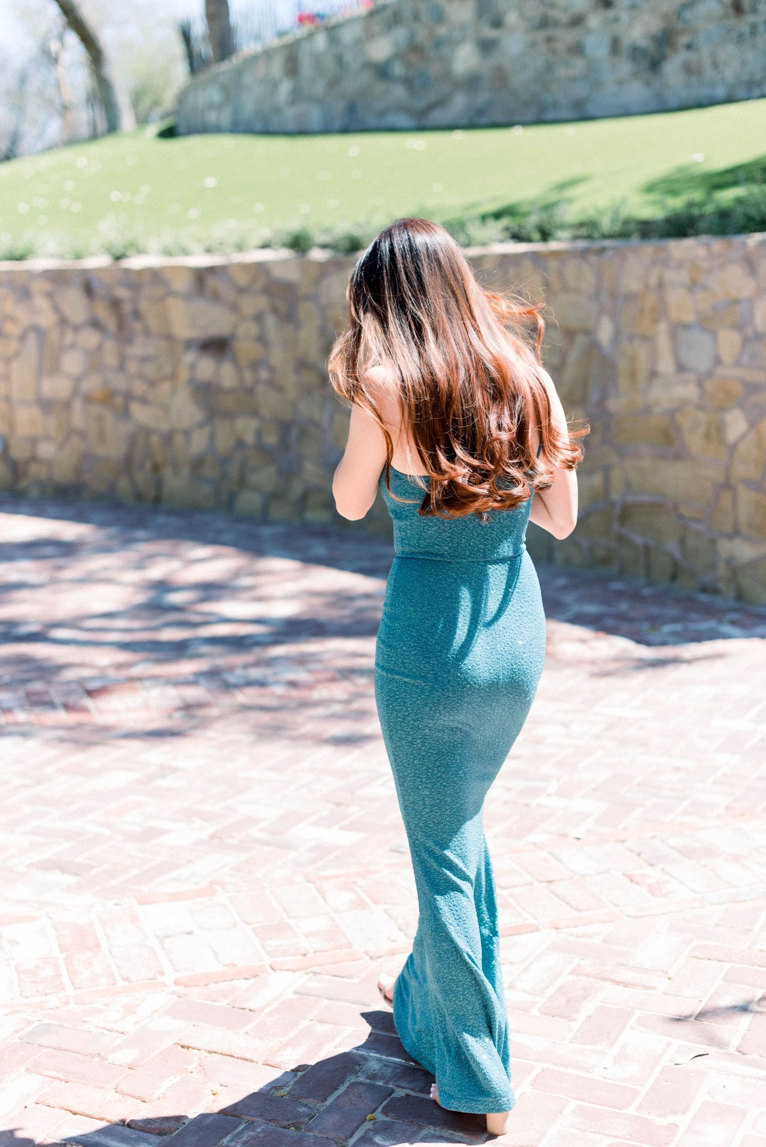 Person in fitted teal gown poses gracefully on brick pathway near stone wall.