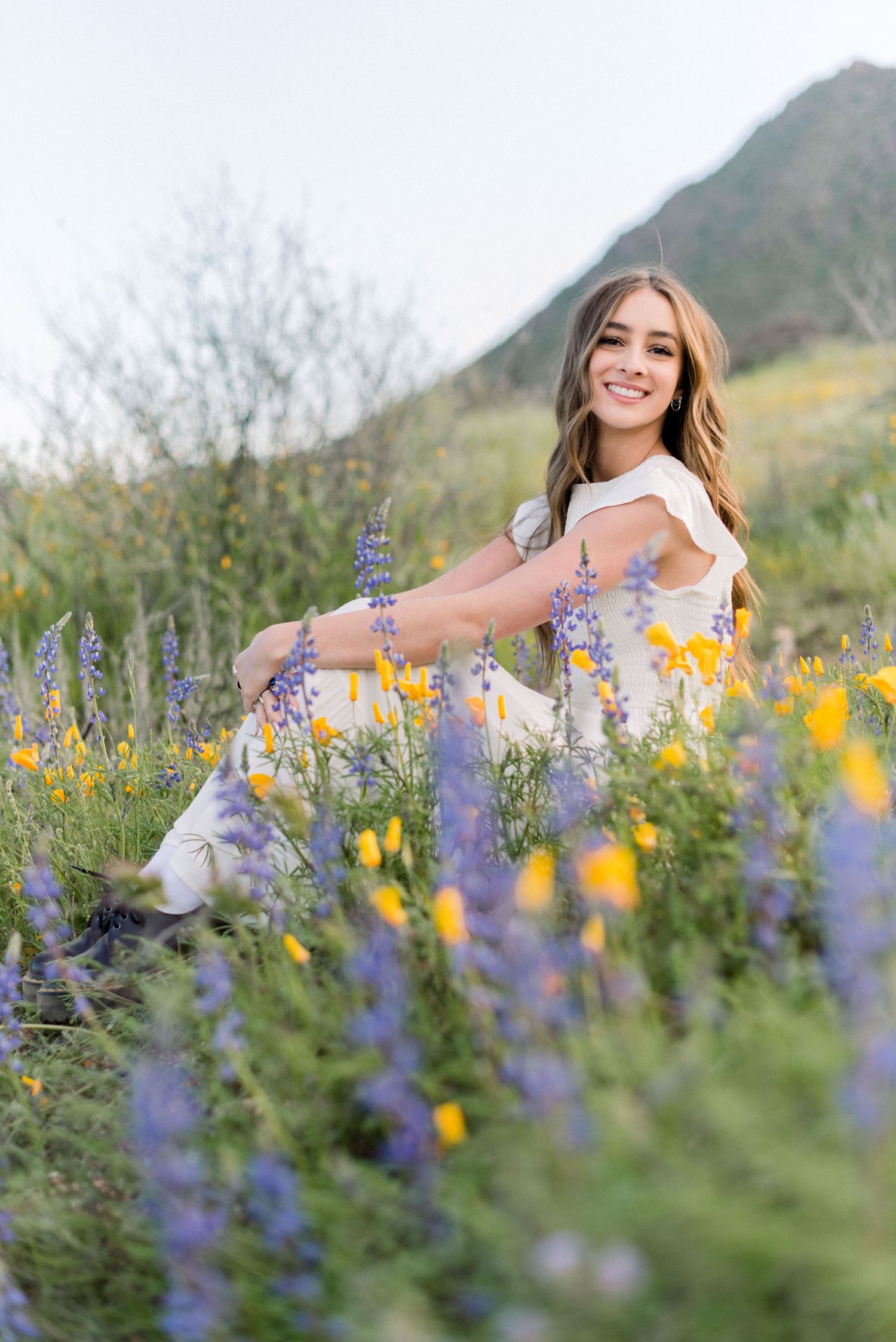 A woman in a white dress poses amid purple lupines and yellow wildflowers on a mountainside during golden hour.
