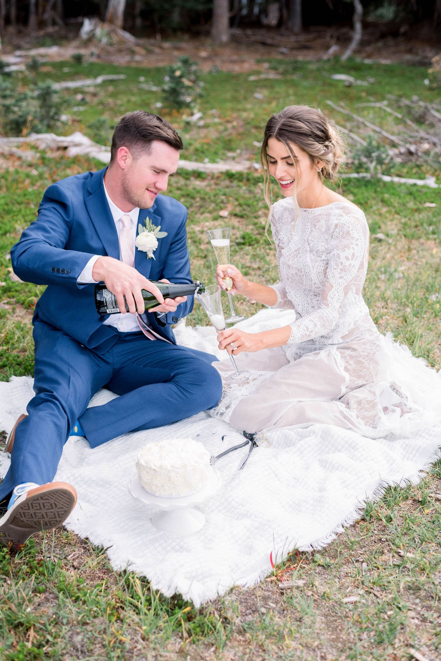 Wedding couple enjoys champagne picnic on white blanket in forest clearing.