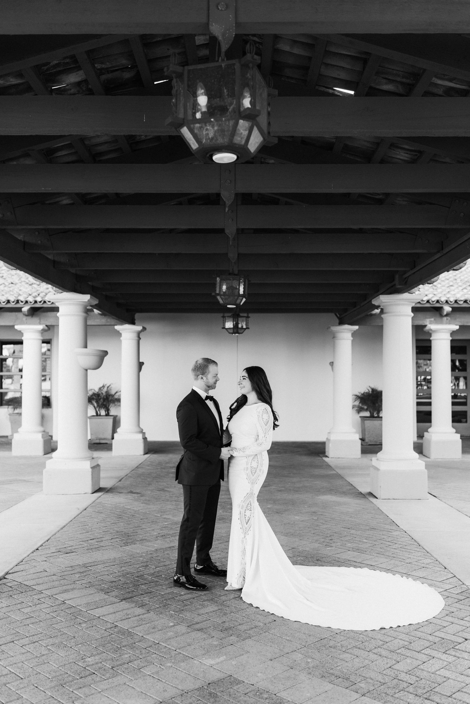 A wedding couple stands intimately under a covered walkway with white pillars and wooden beams in black and white.