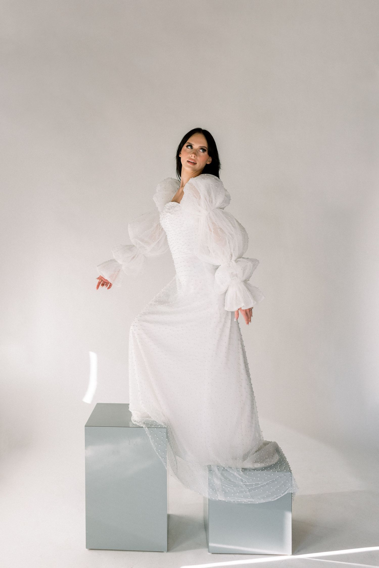 Woman in dramatic white dress poses on silver cube pedestals against neutral backdrop.