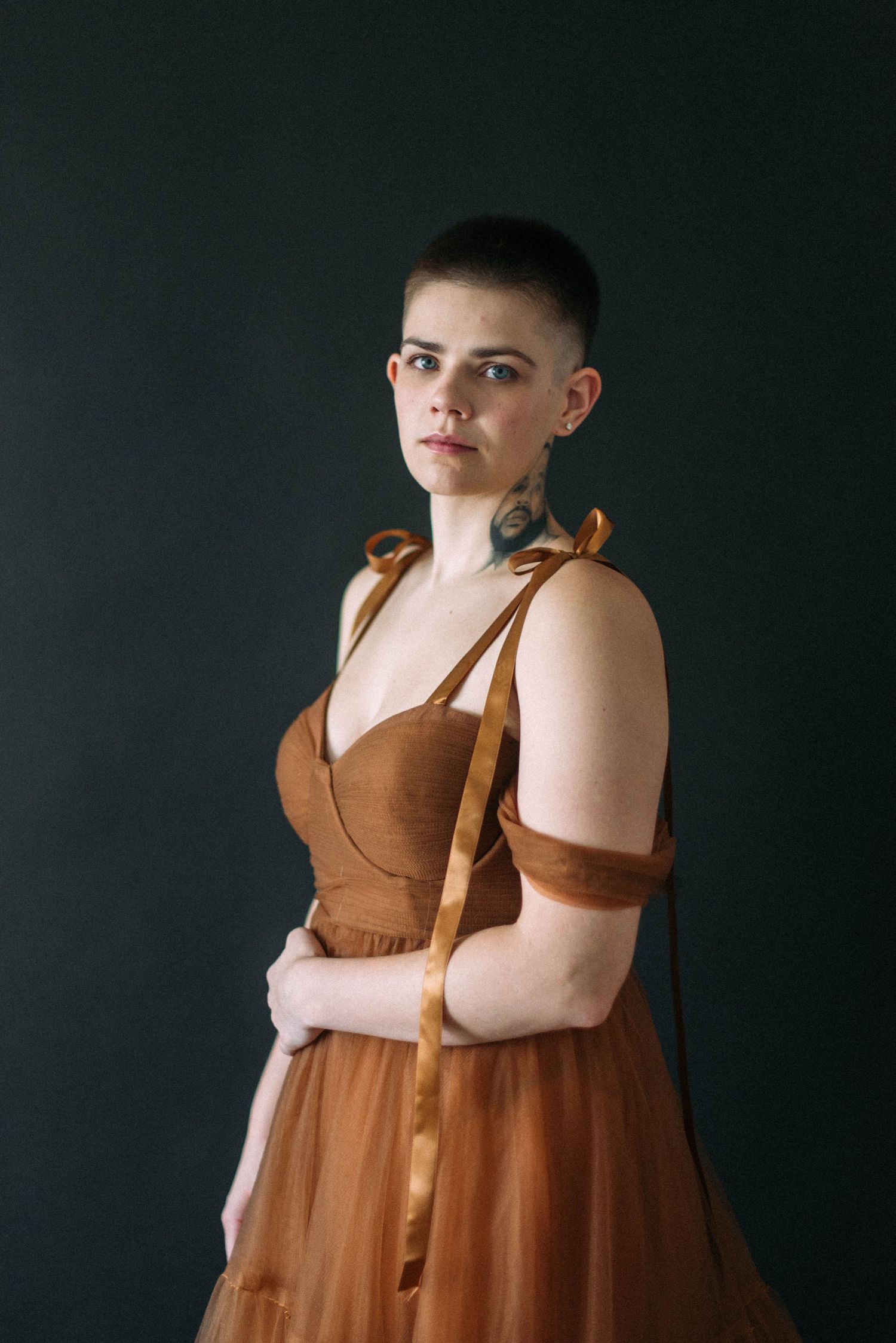 Person in a brown formal dress with ribbon straps posing against dark backdrop.