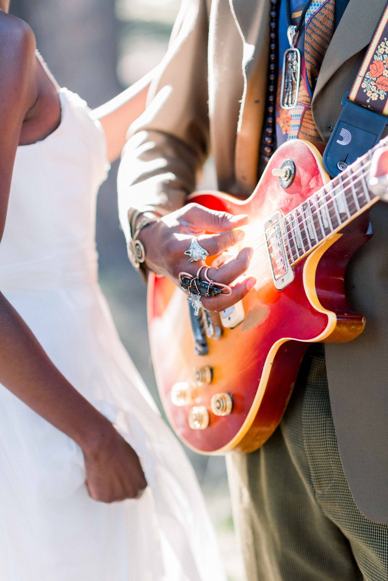 A detail shot of a sunburst electric guitar held by a groom in tan suit next to a white wedding dress.
