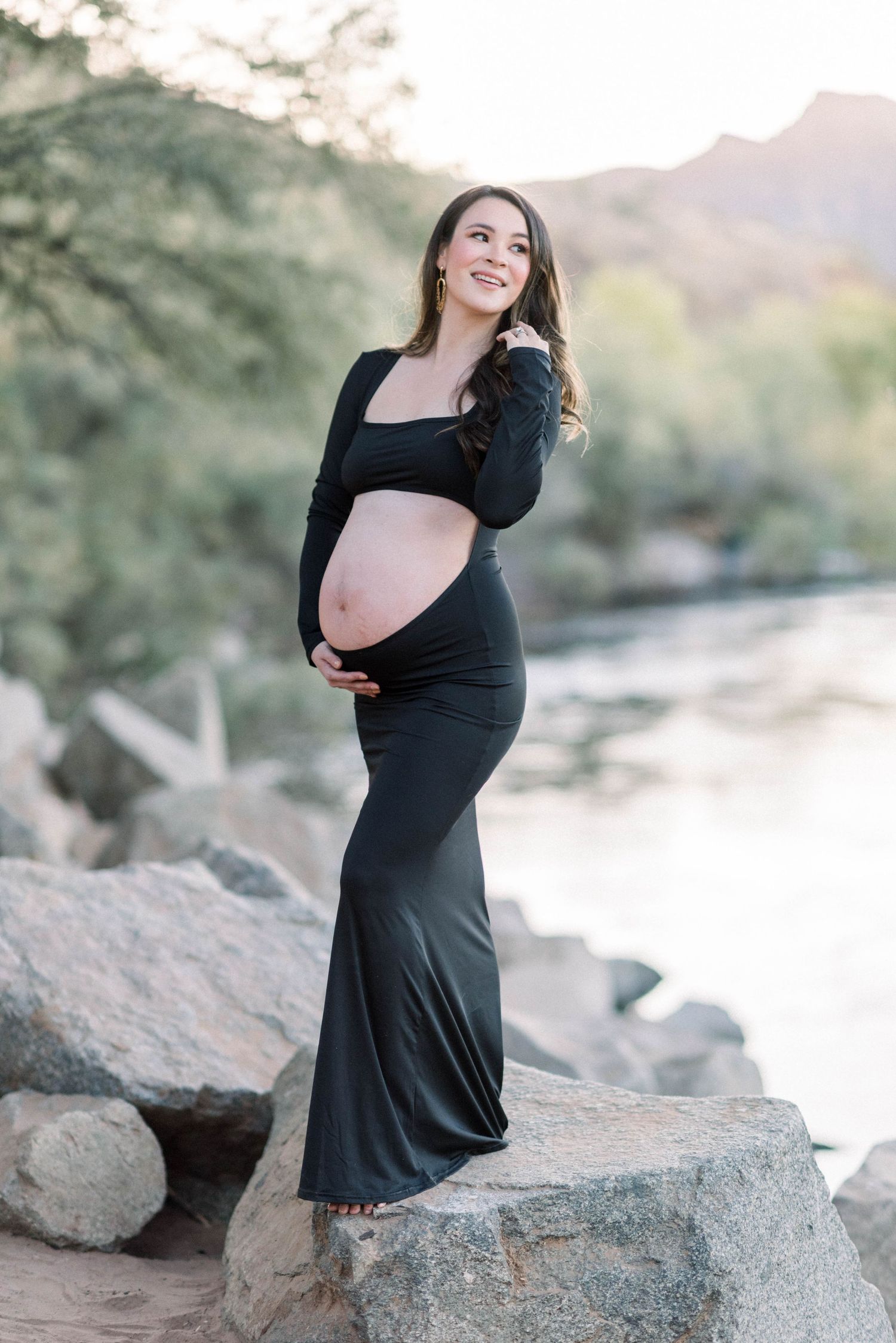 A woman in a black dress poses on rocky terrain during a maternity photoshoot at sunset.