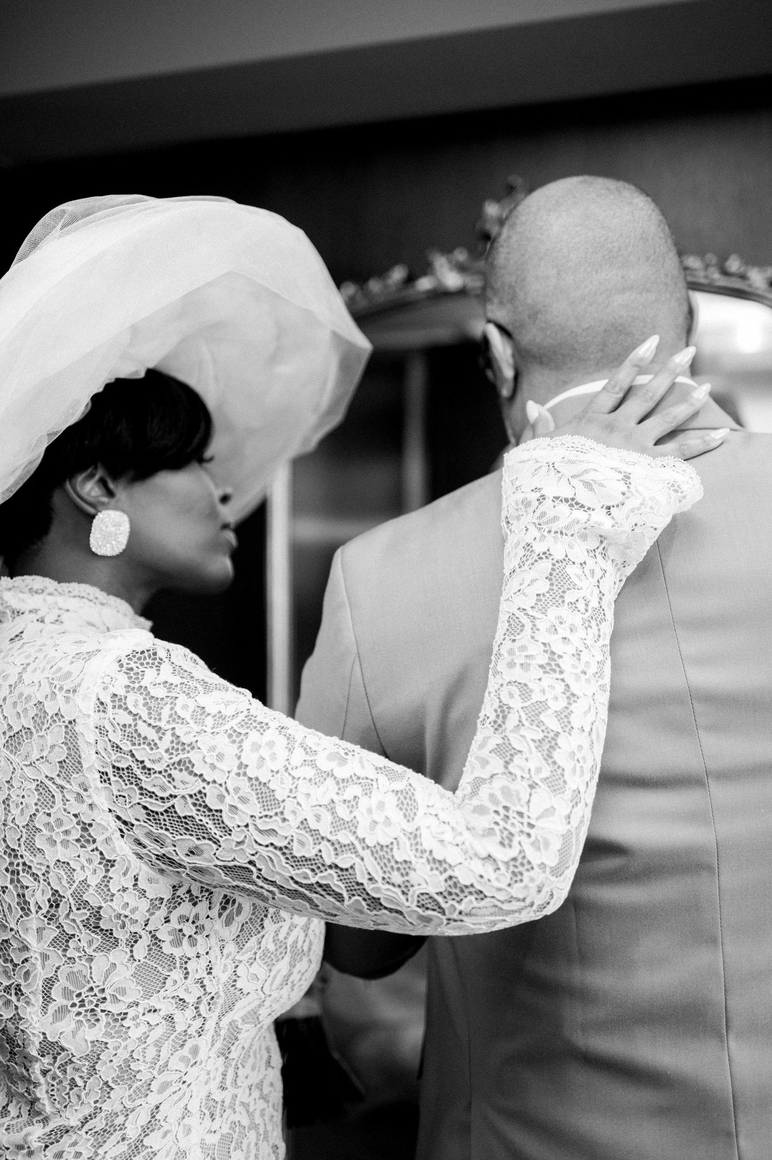 Black and white wedding photo showing tender moment between couple in lace and formal attire.