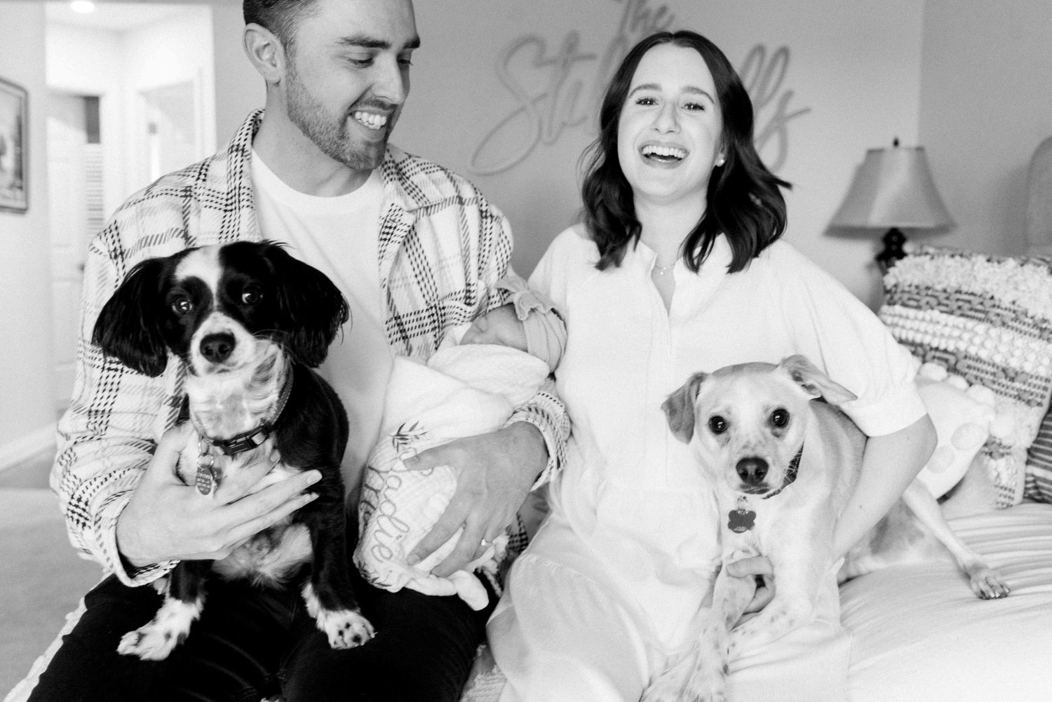 Black and white family photo with newborn baby and two dogs on couch.