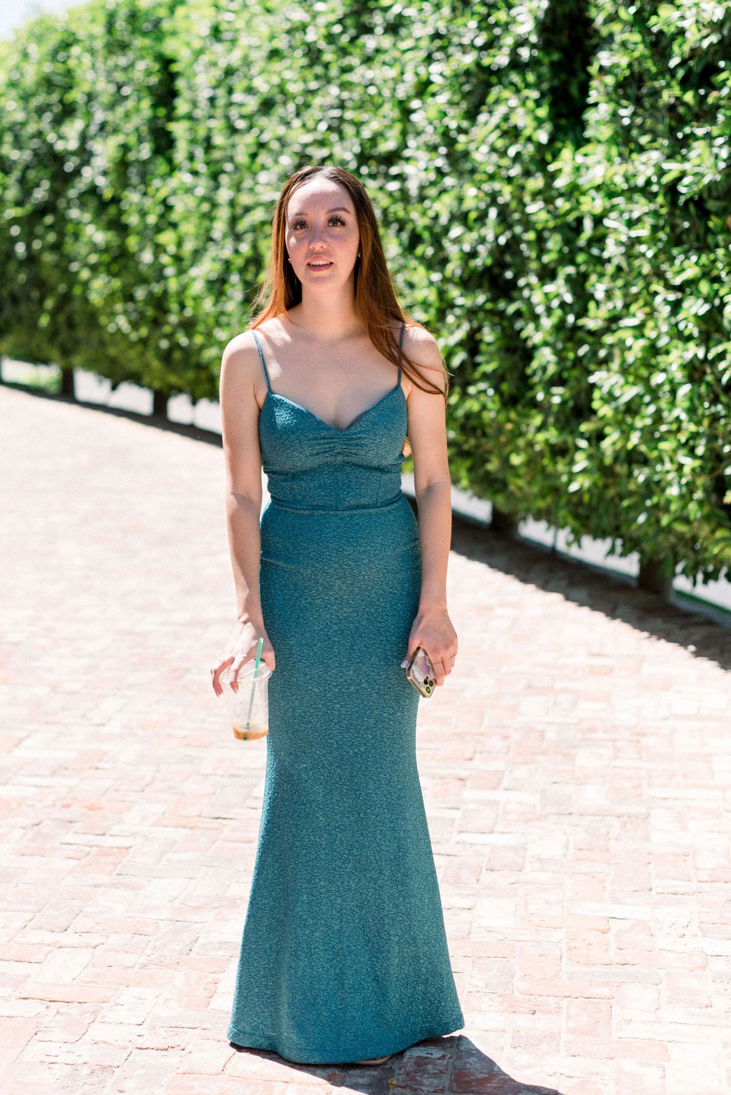 Woman in a teal sparkly formal dress poses in front of a green hedge wall.