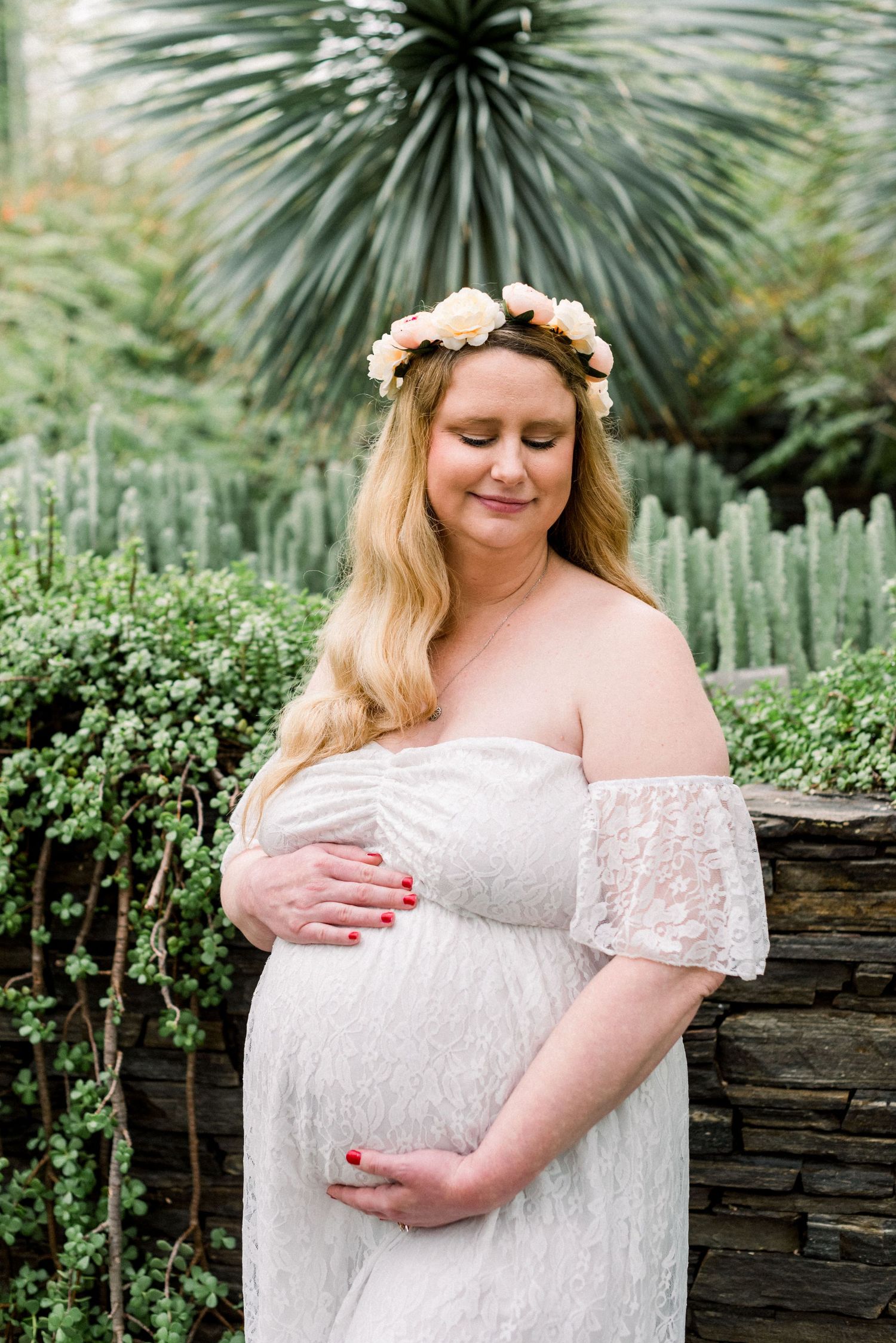 Maternity portrait wearing a white lace dress and floral crown against a garden backdrop with succulents.