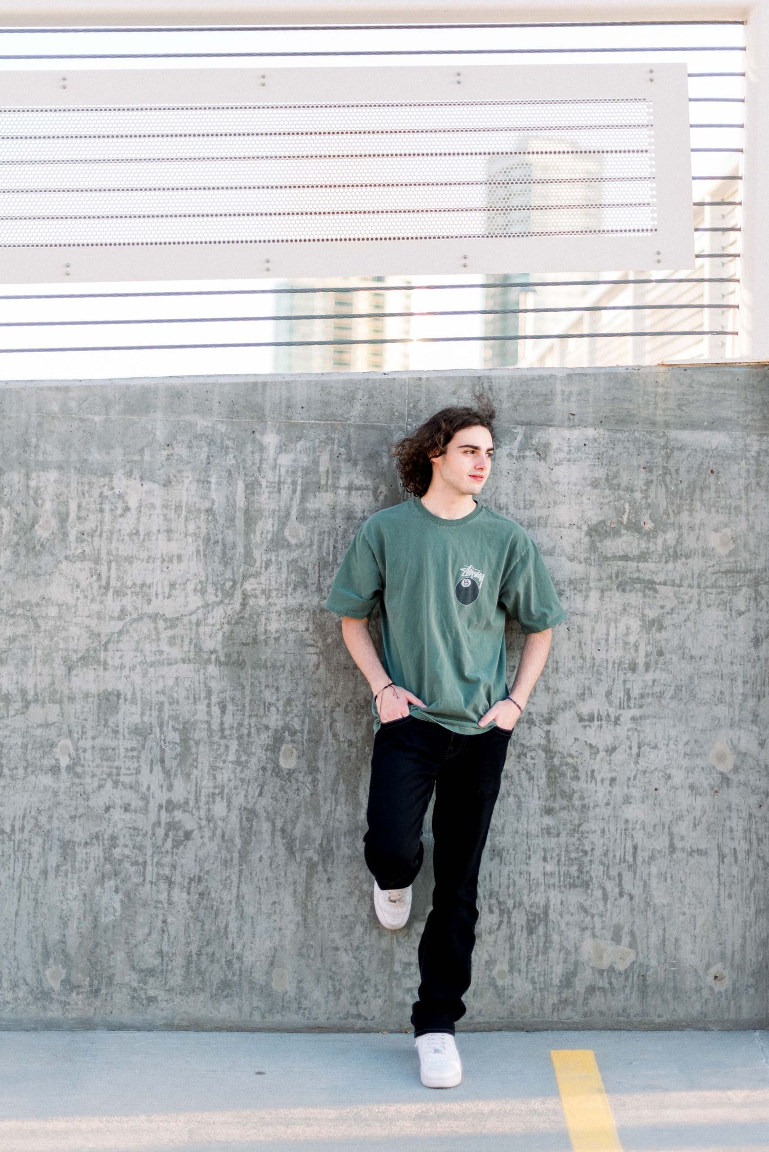Person in green t-shirt leans against concrete wall in casual pose with hands in pockets.
