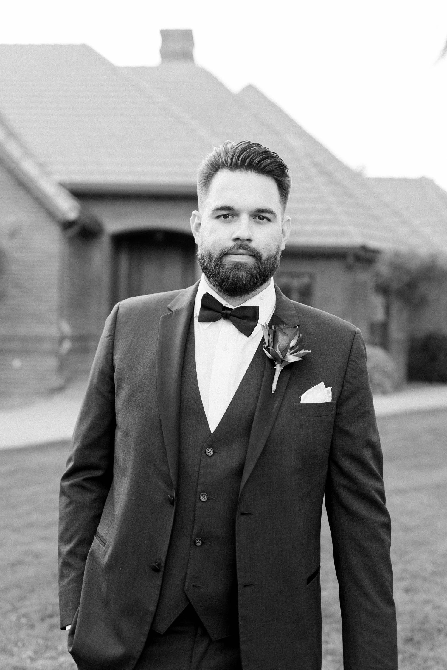 Close-up portrait of groom in classic tuxedo with boutonniere against brick building backdrop.