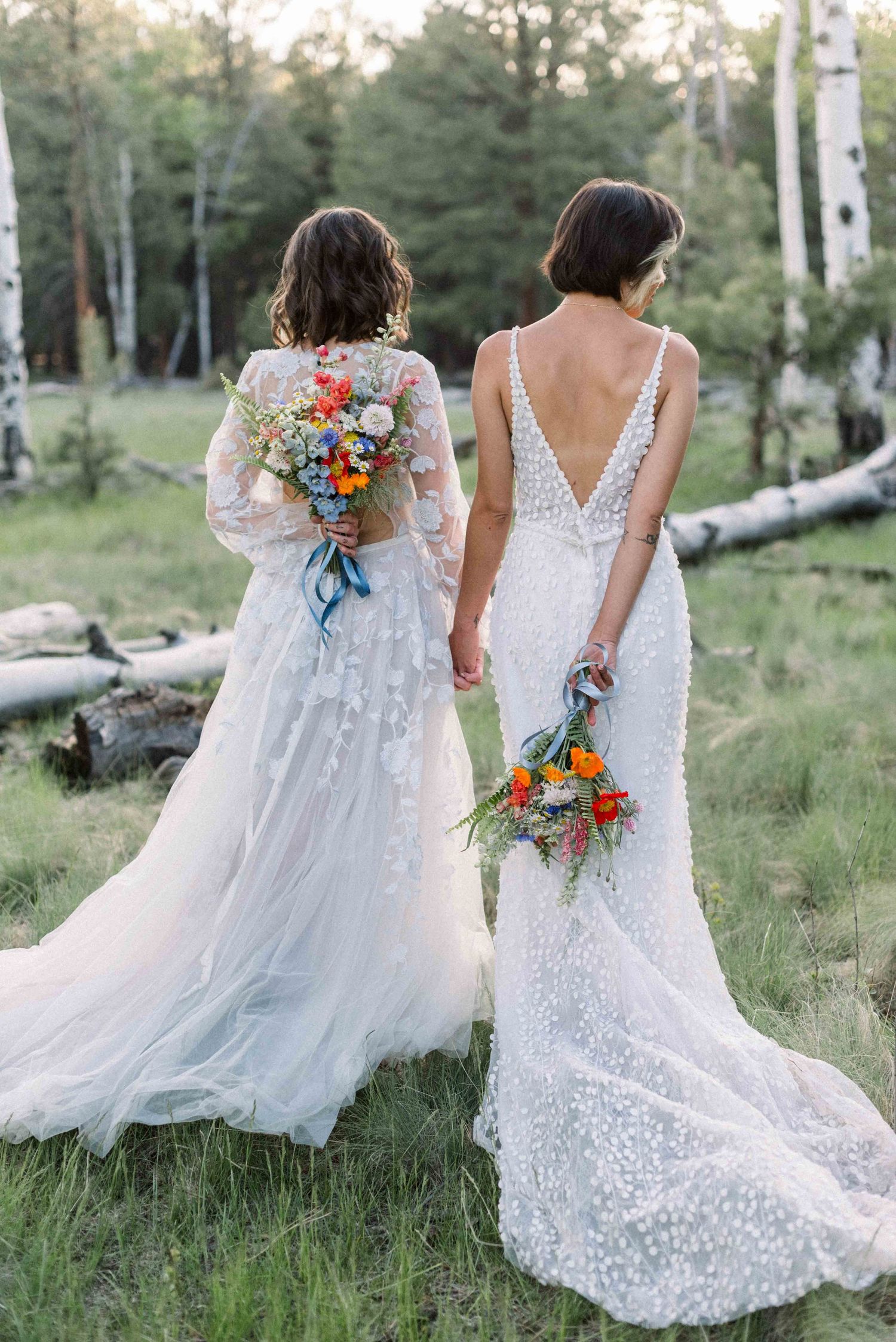 Two brides in white gowns holding vibrant bouquets against aspen grove backdrop.