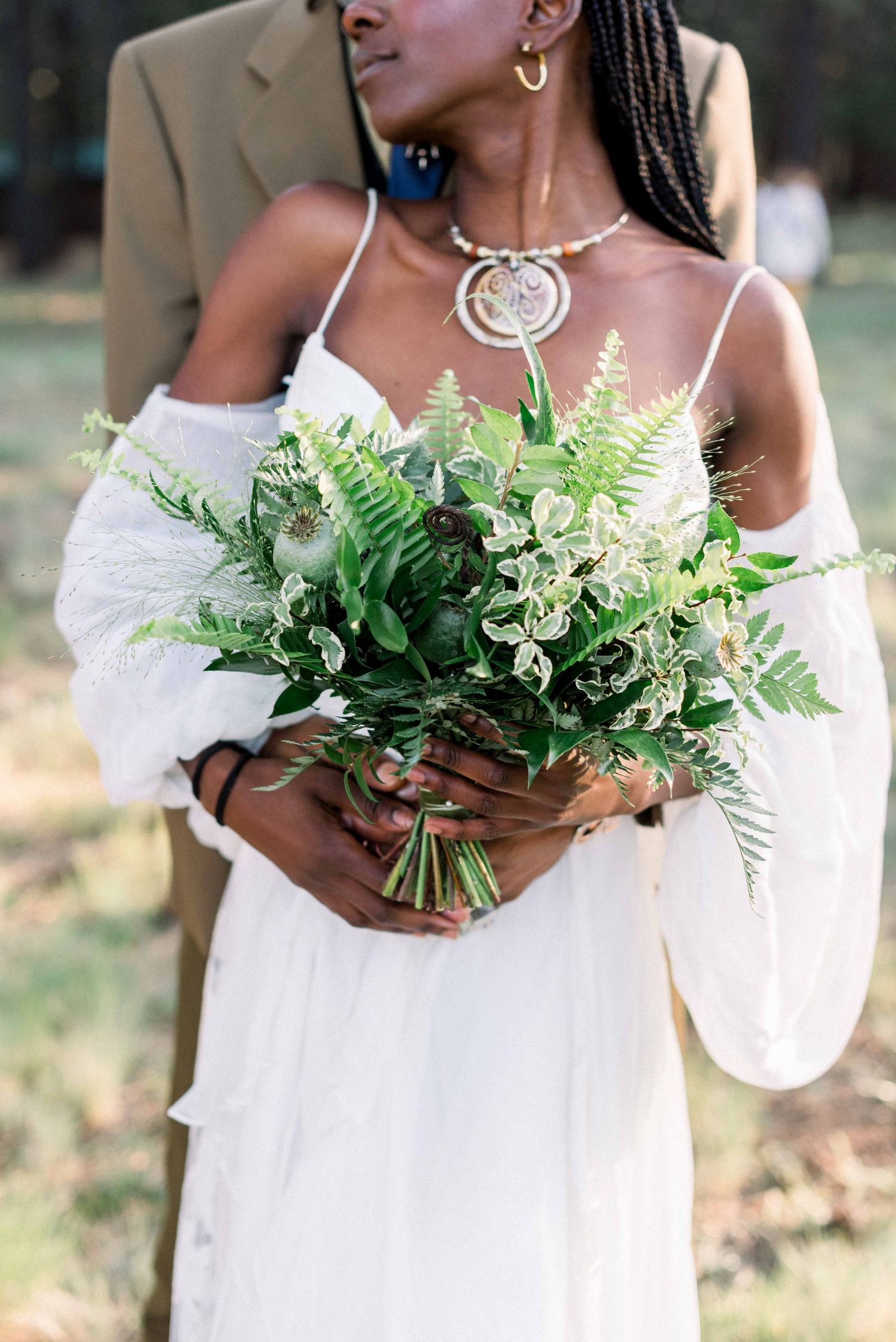 Elegant bride in white dress holds a lush bouquet of ferns and greenery while wearing a statement necklace.