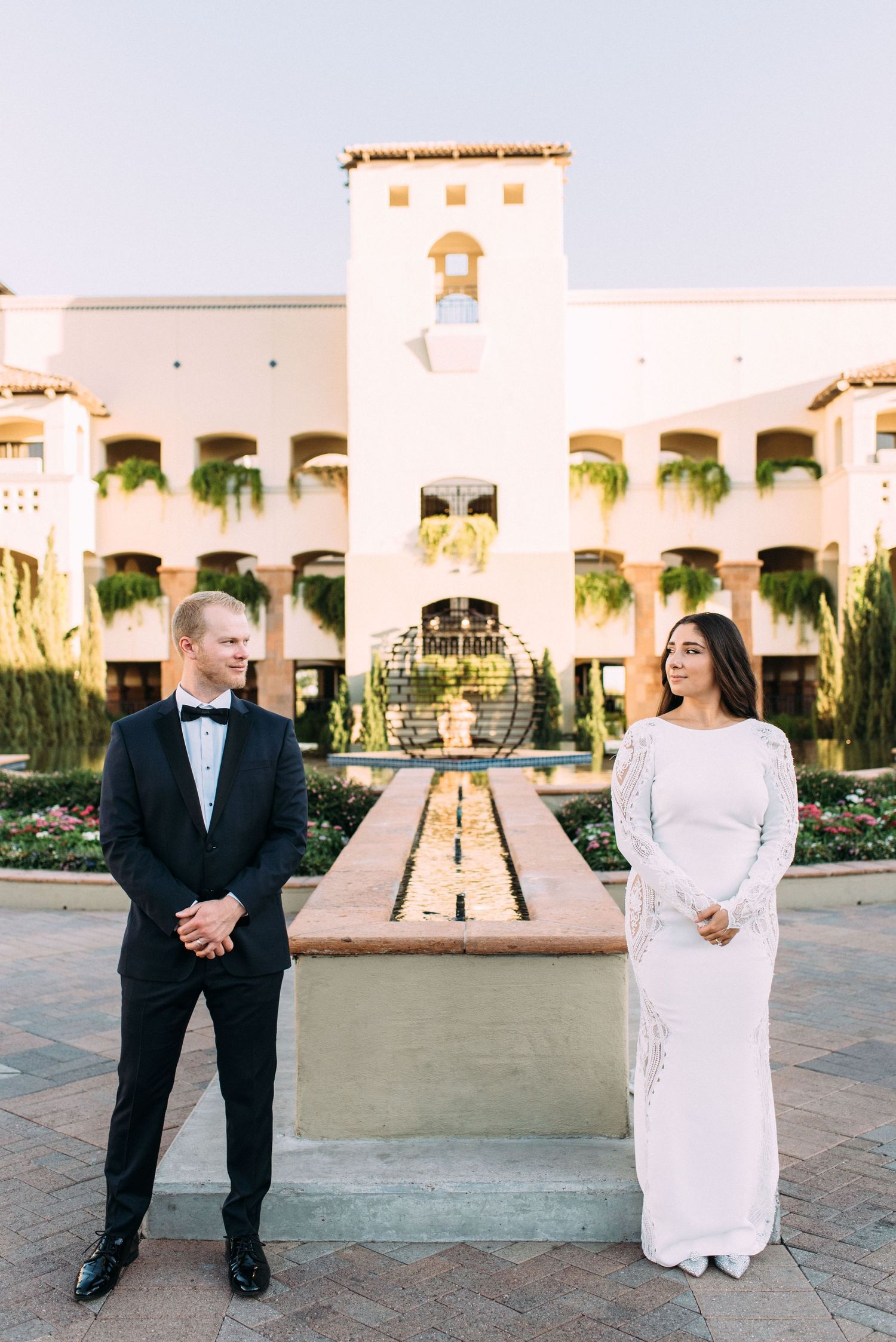 A wedding couple poses formally in front of a Mediterranean-style building with fountain.
