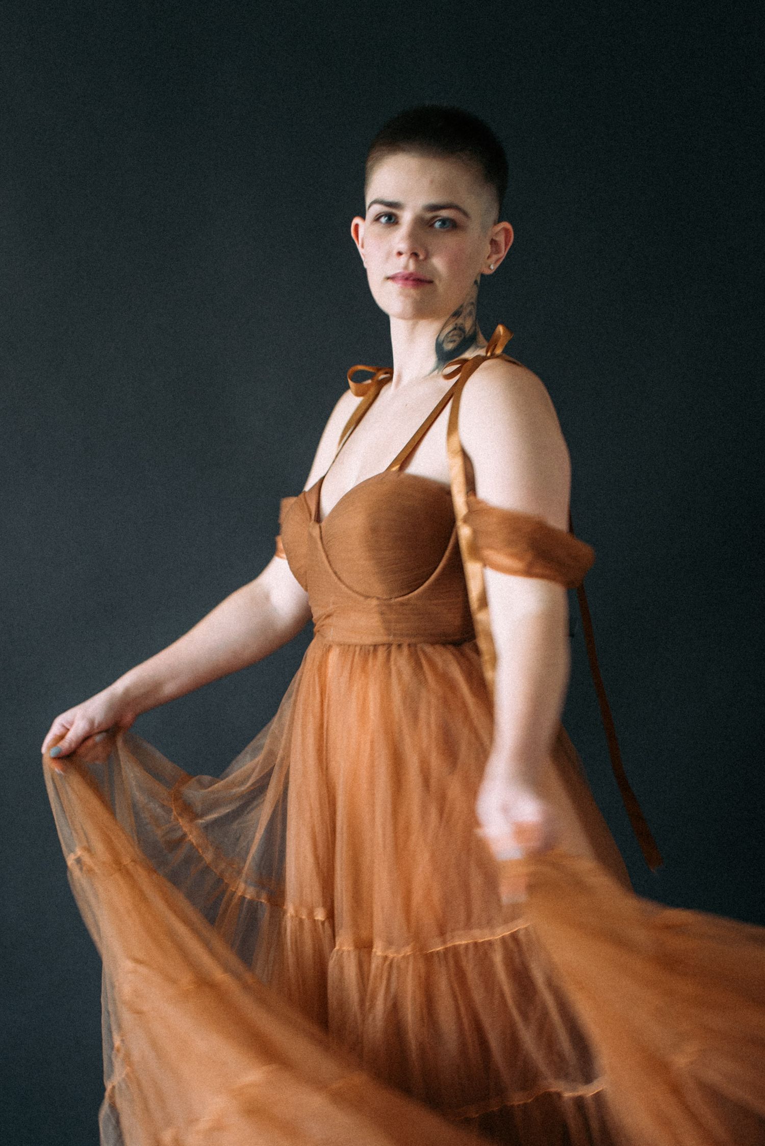 A model in a flowy brown tulle dress poses against a dark background while holding the dress skirt.