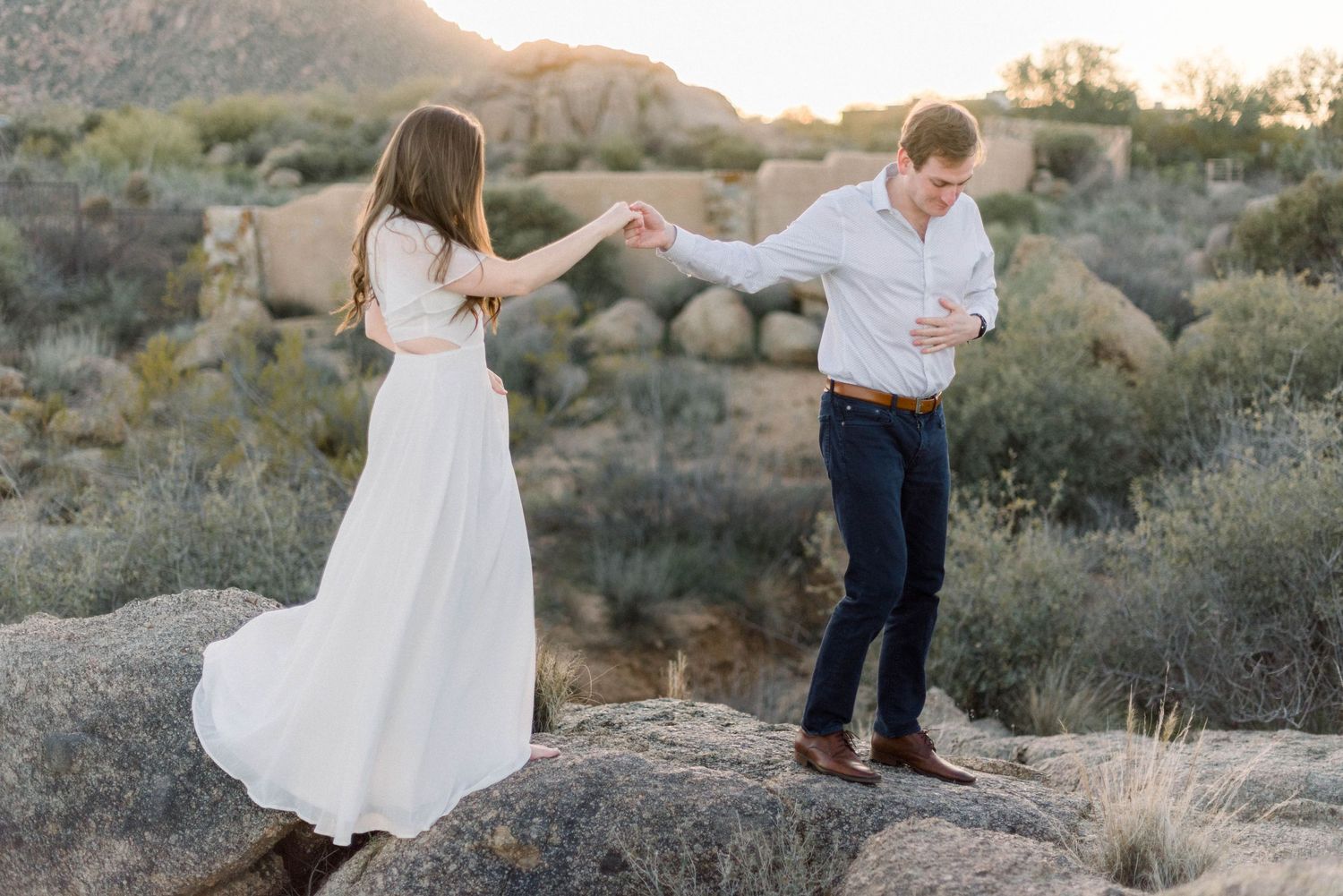 Playful couple in white and blue outfits dancing on desert rocks at sunset.