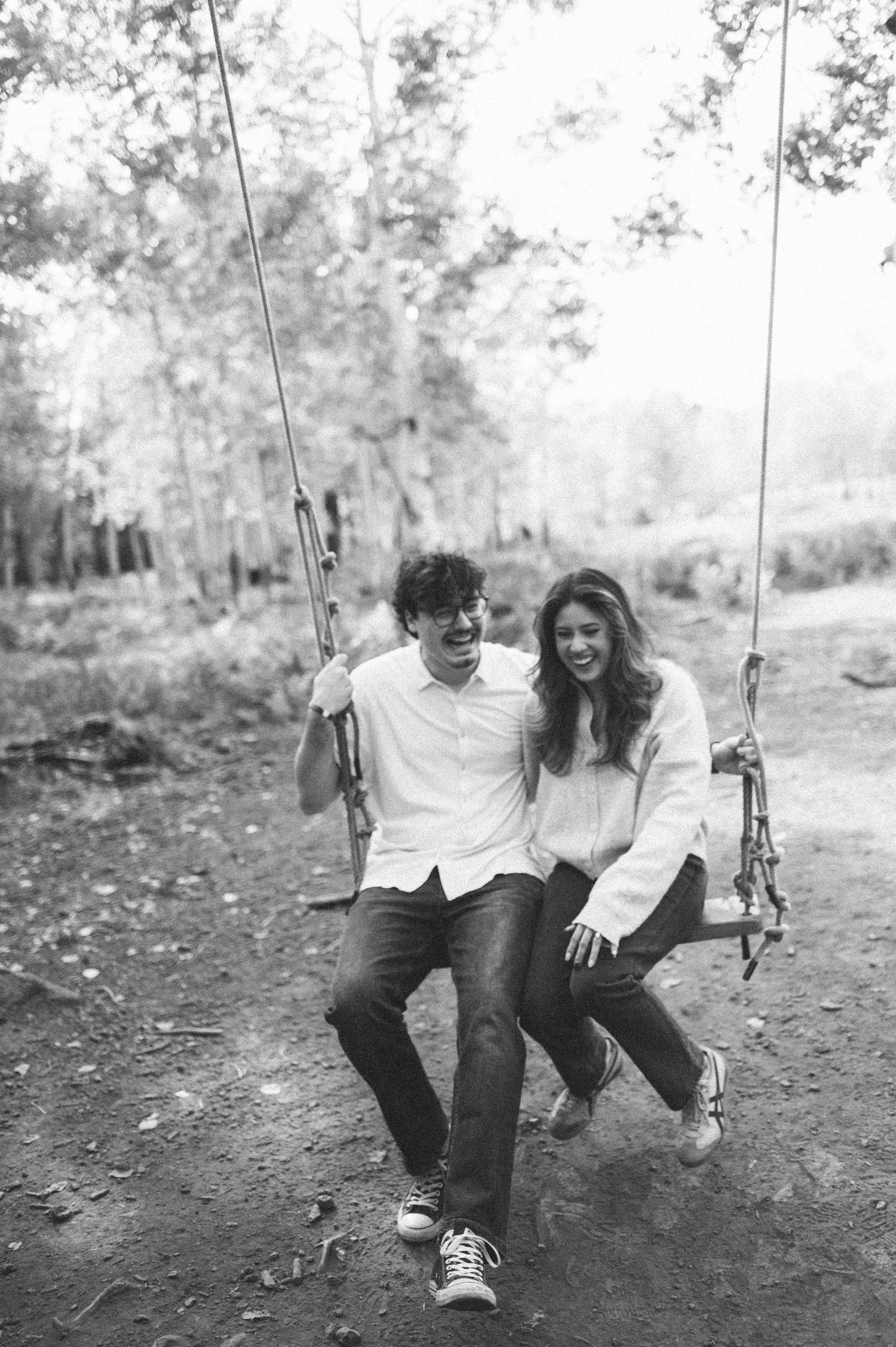 A joyful couple shares a swing together in a black and white photo taken in a wooded outdoor setting.