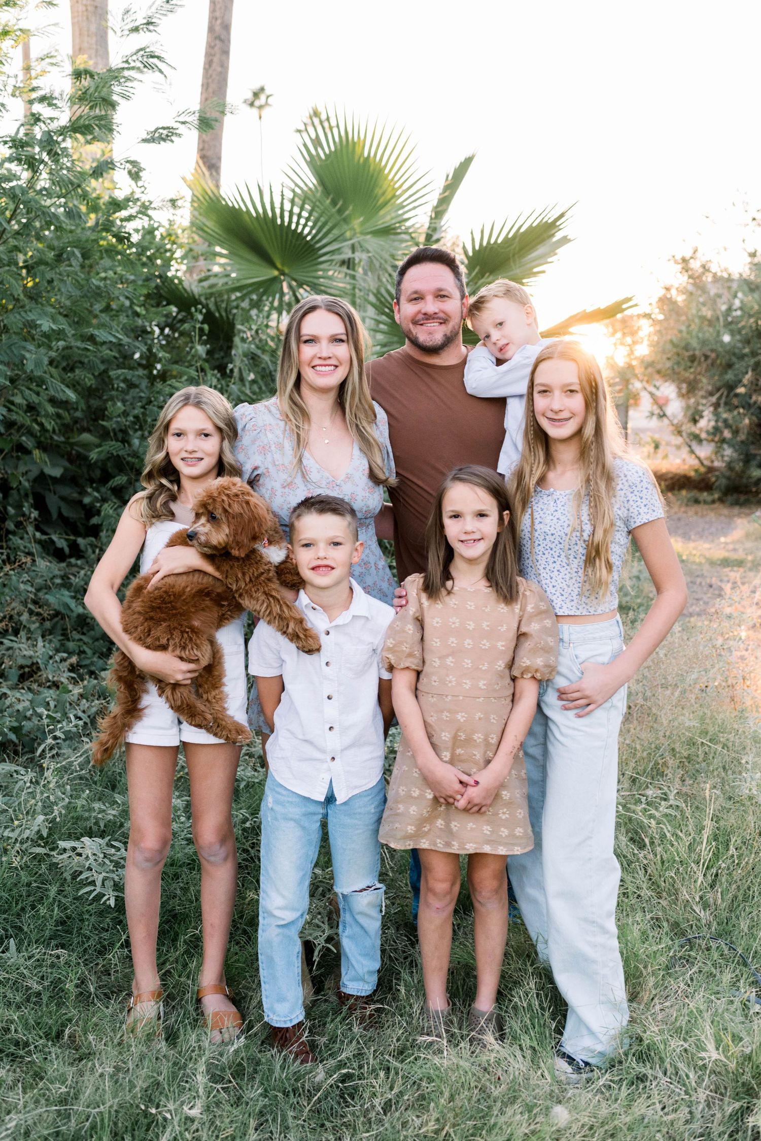 Family wearing coordinated neutral outfits poses together outdoors during golden hour with their small brown dog.